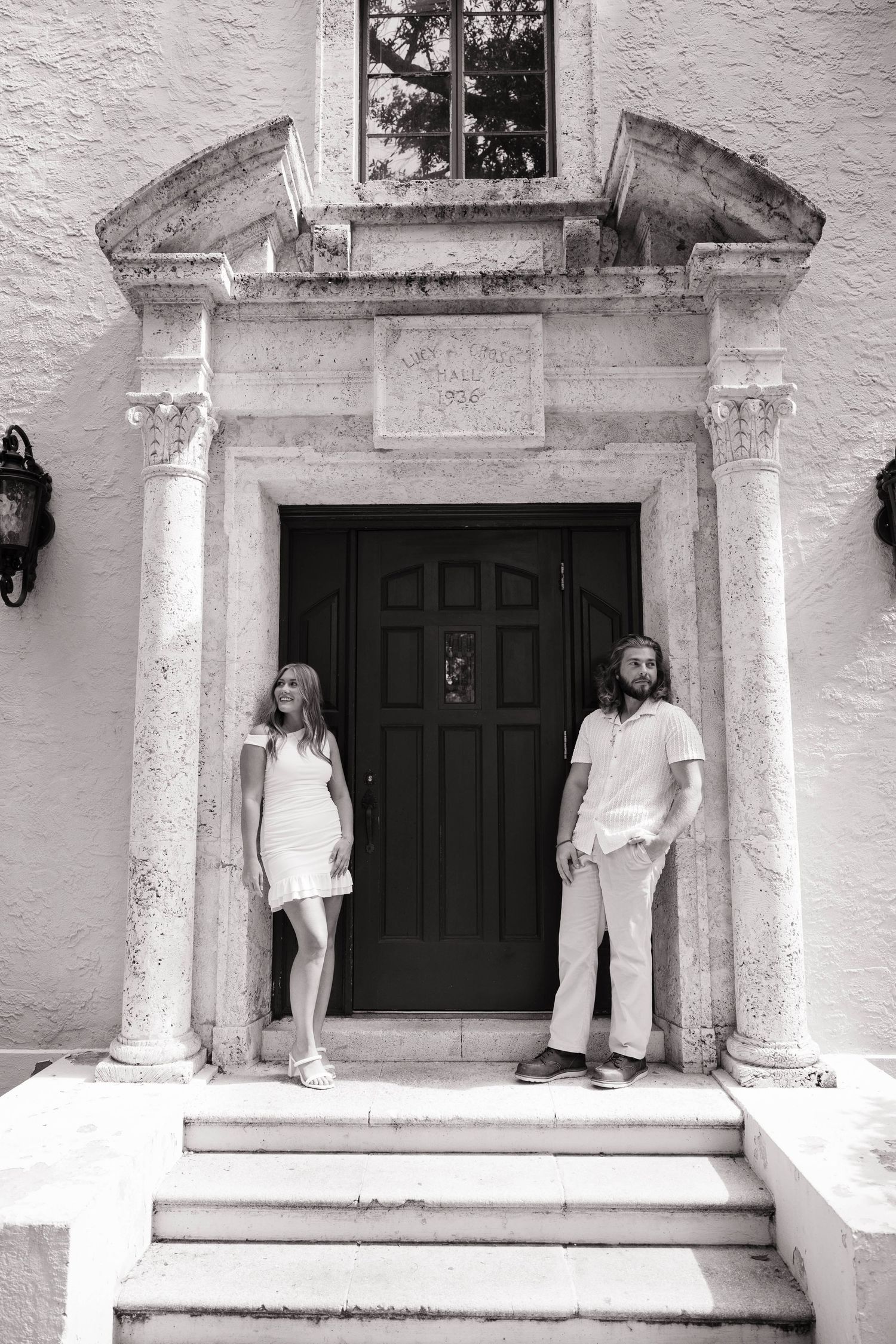 Black and white photo of couple standing on steps of historic stone doorway entrance of elegant building.