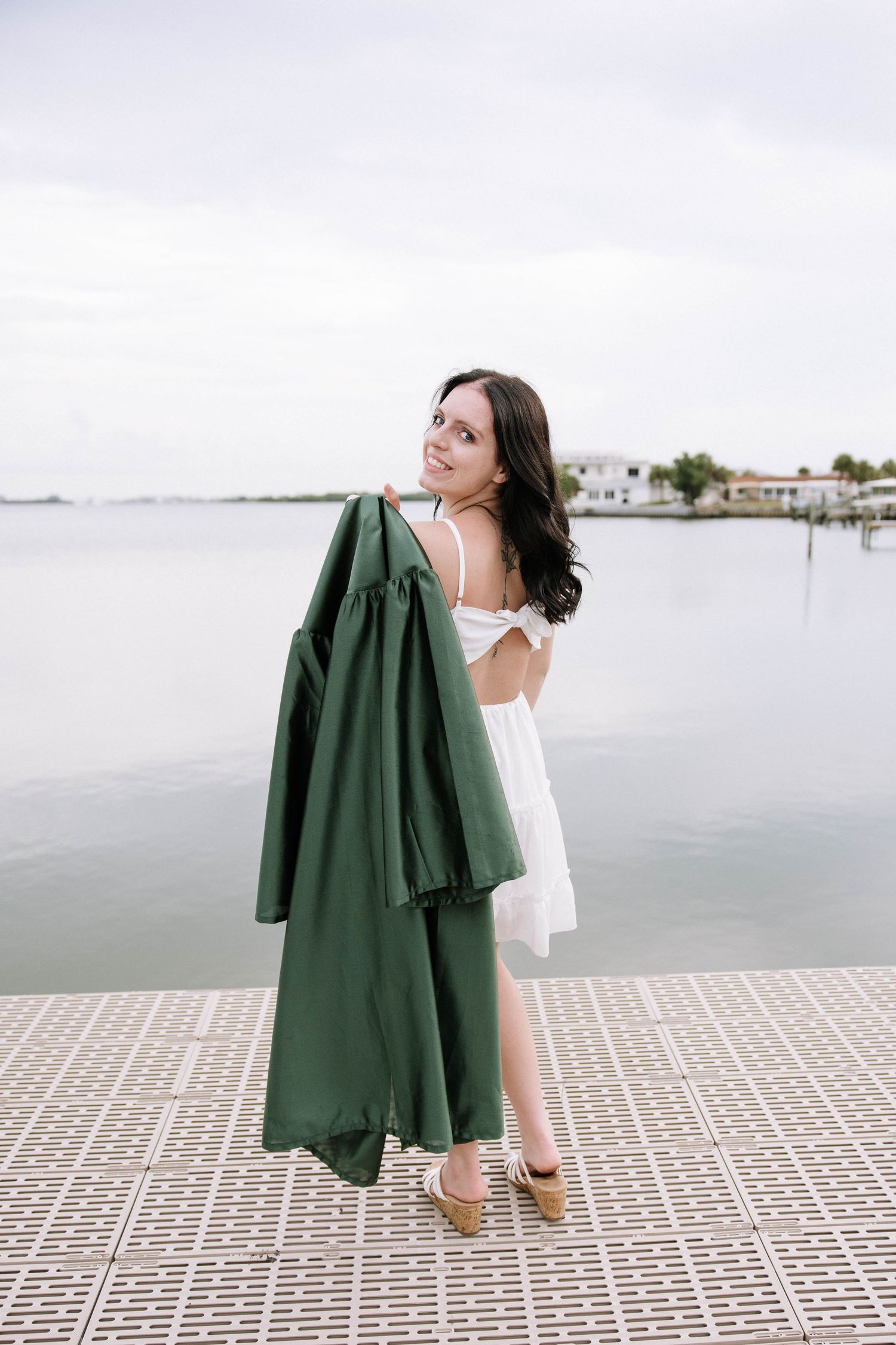 Smiling brunette woman in white dress holding green graduation gown on waterfront dock with marina behind her.Photographers at Anna Maria Is