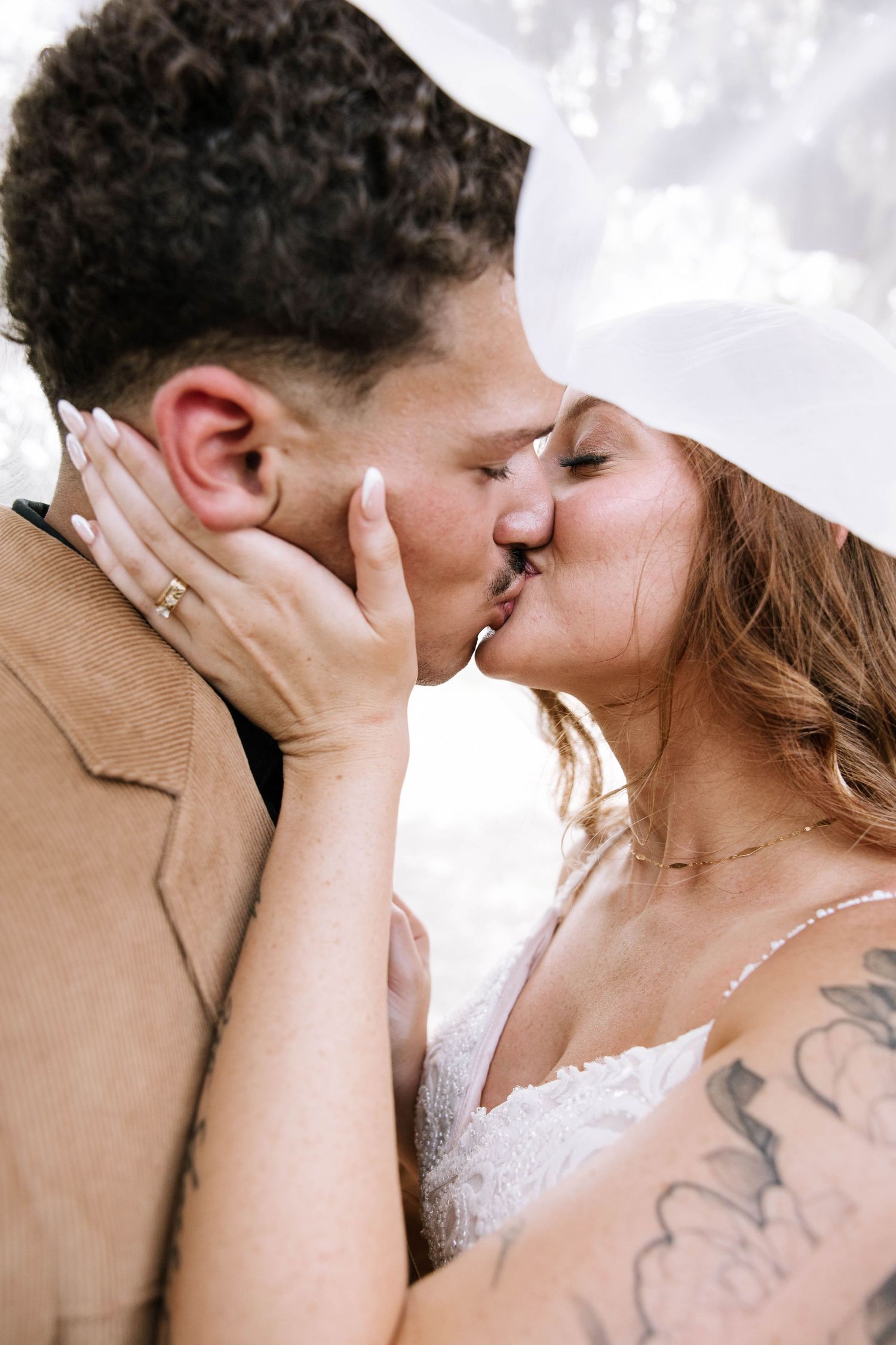 Close-up of tattooed bride and curly-haired groom sharing tender kiss under wedding veil.