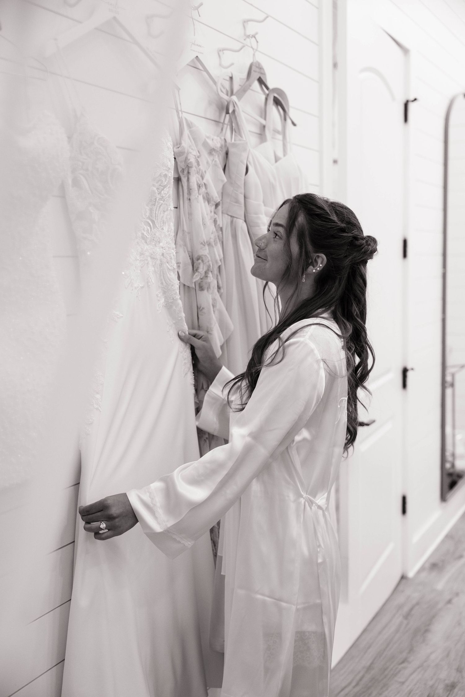 A bride in a white satin robe smiles while looking at wedding dresses hanging on a shiplap wall.