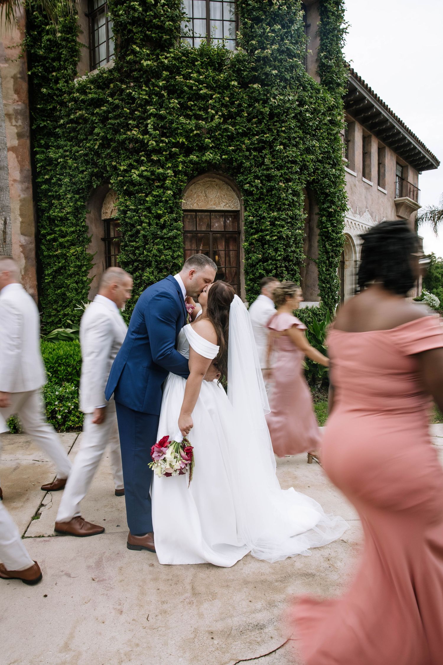 Bride and groom share a kiss outside an ivy-covered stone building while wedding party walks by.