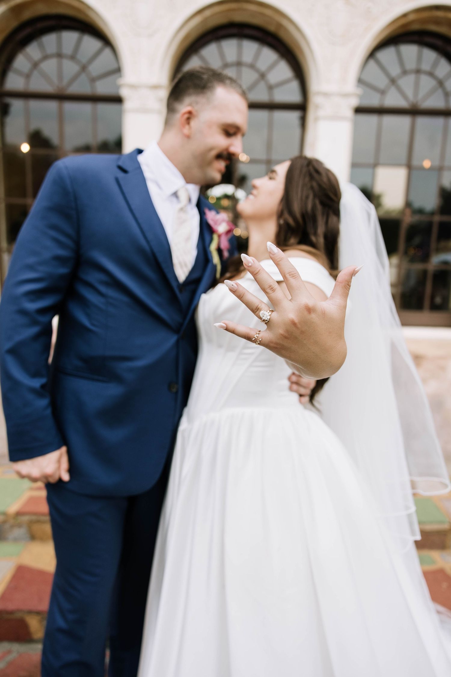 Bride and groom sharing a romantic moment, showing off wedding rings, groom in navy suit, bride in white gown with veil.
