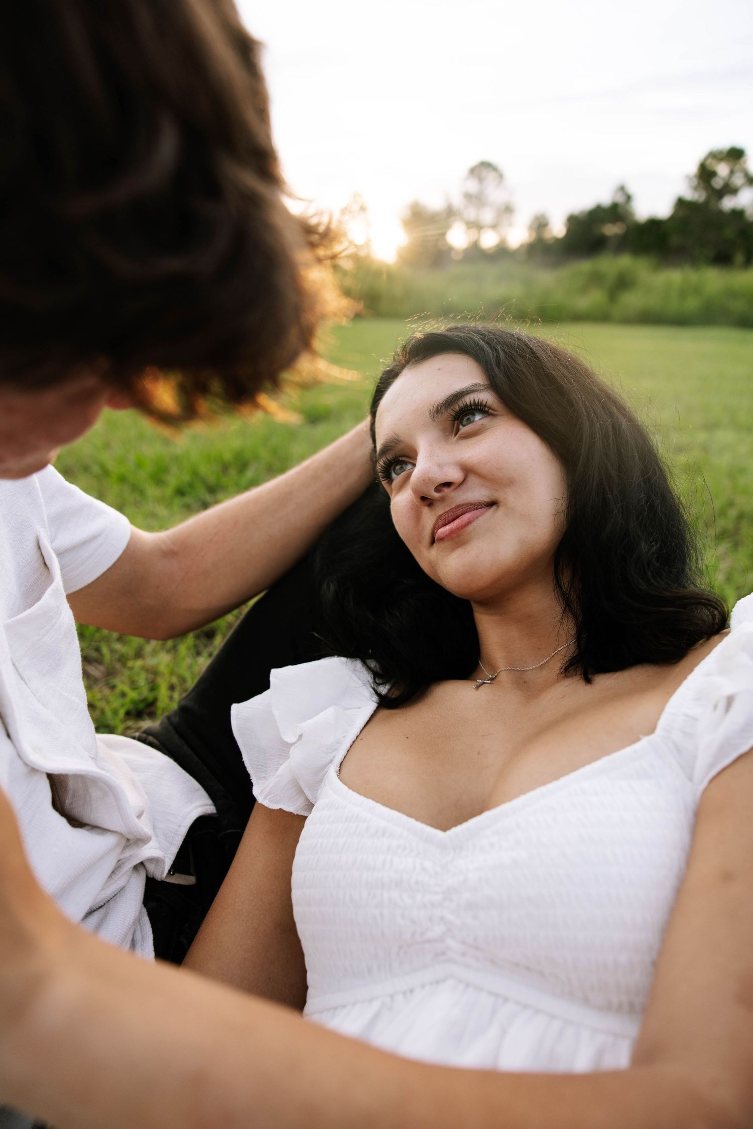 A young woman in a white dress smiles up at someone touching her hair during a golden hour sunset outdoors.