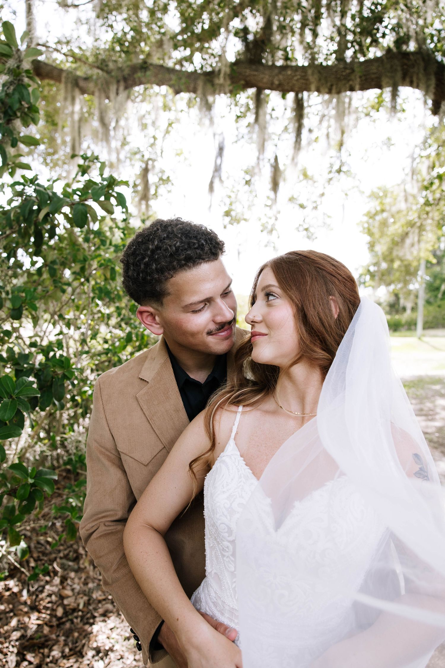 Bride and groom gazing lovingly at each other under Spanish moss-draped trees in an outdoor garden setting.