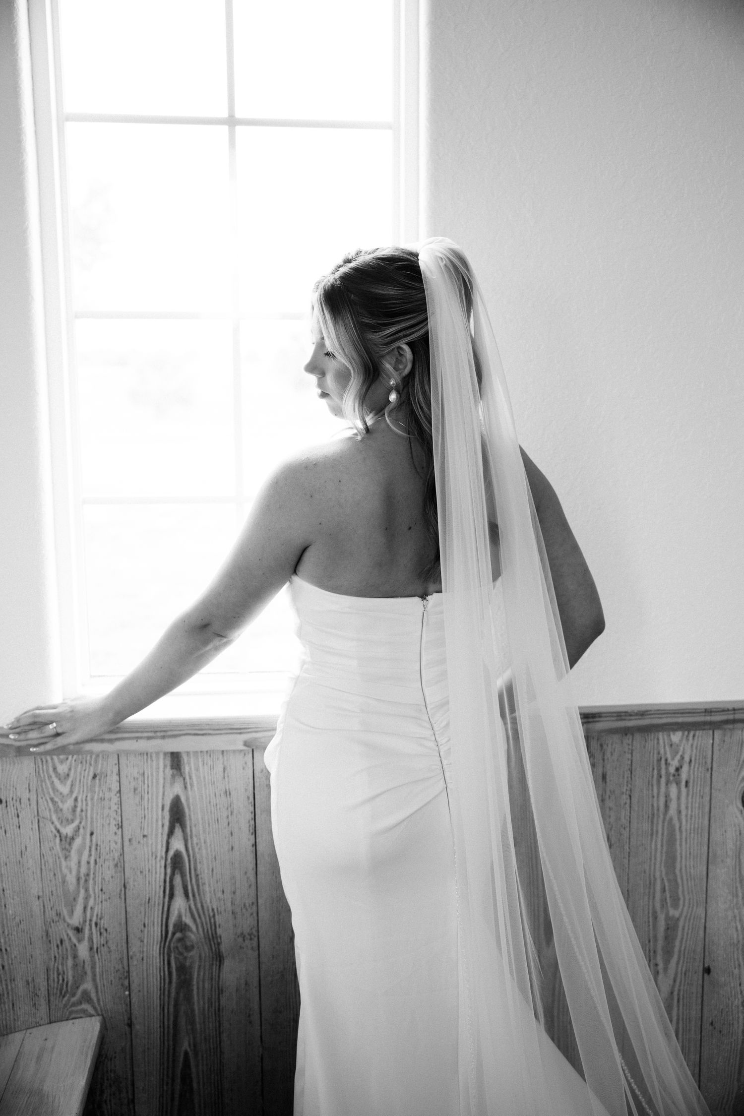 Elegant black and white photo of bride in fitted gown and long veil standing by chapel window.