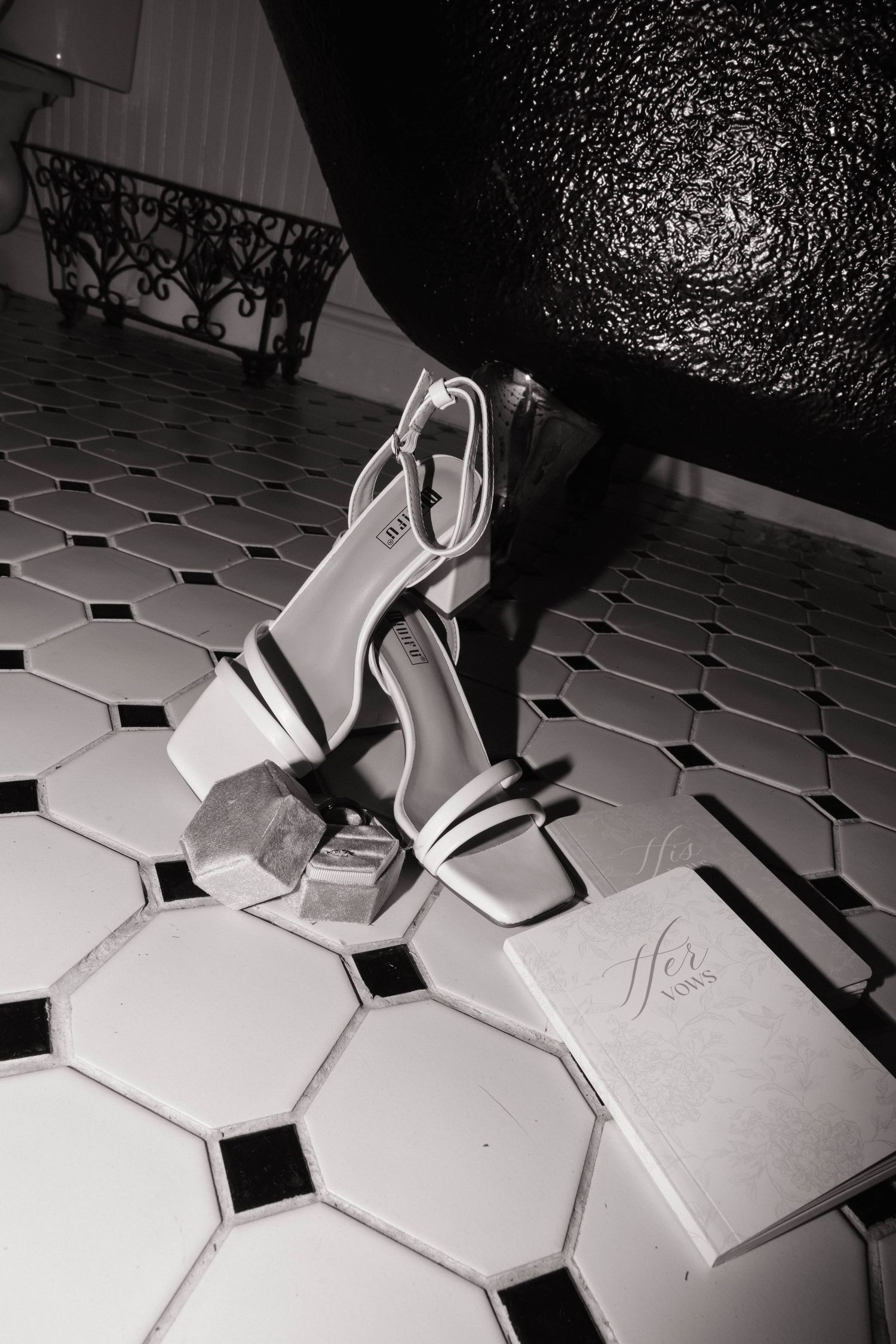 A black and white photo of white strappy heeled sandals resting on hexagonal tile flooring beside a box.