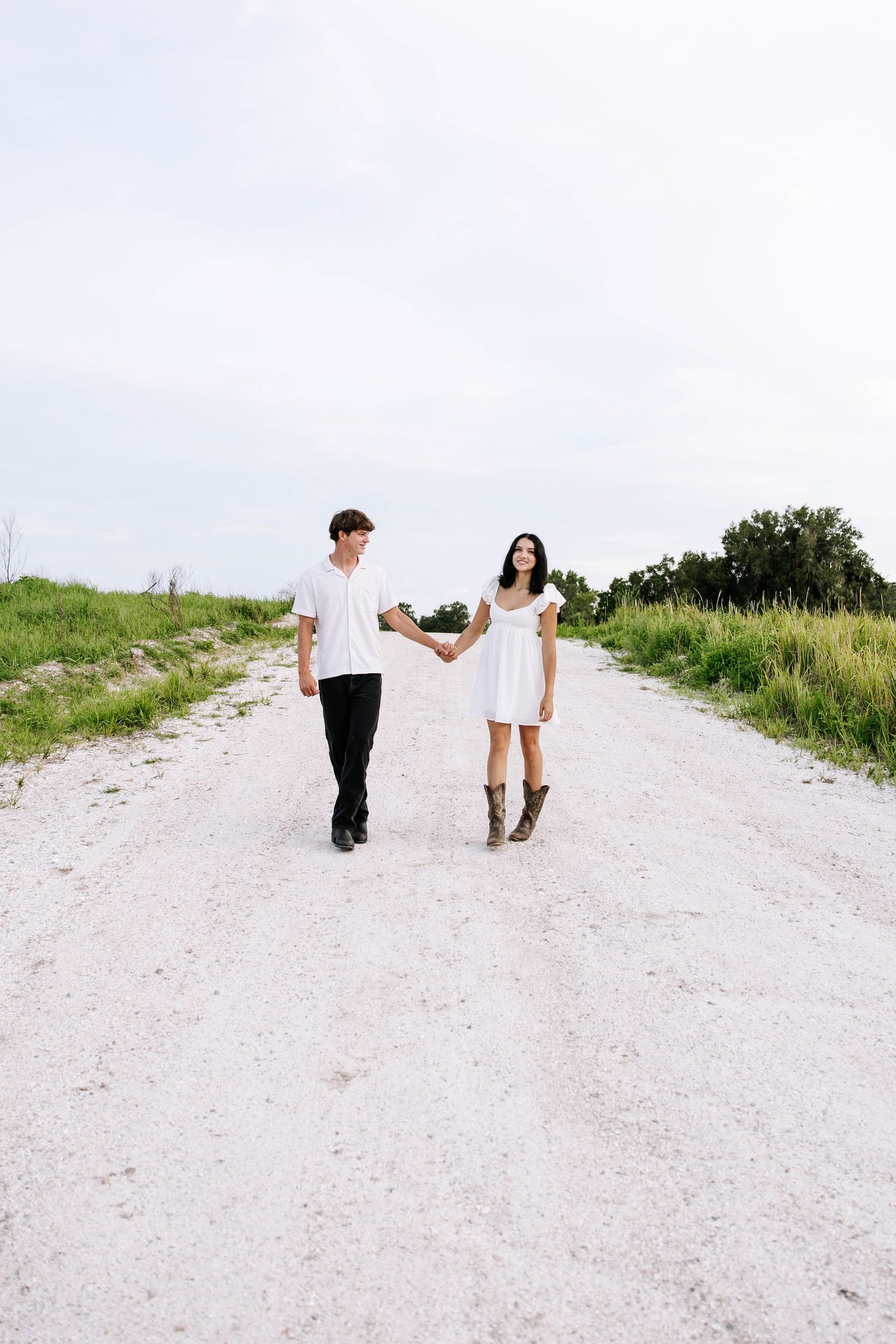 A young couple holding hands while walking on a white gravel road surrounded by green fields under an overcast sky.