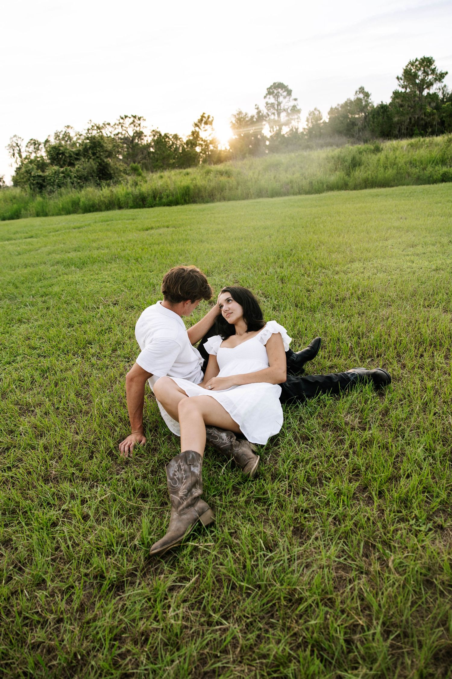 Couple in white outfits sitting on green grass field at sunset, sharing a romantic moment outdoors.