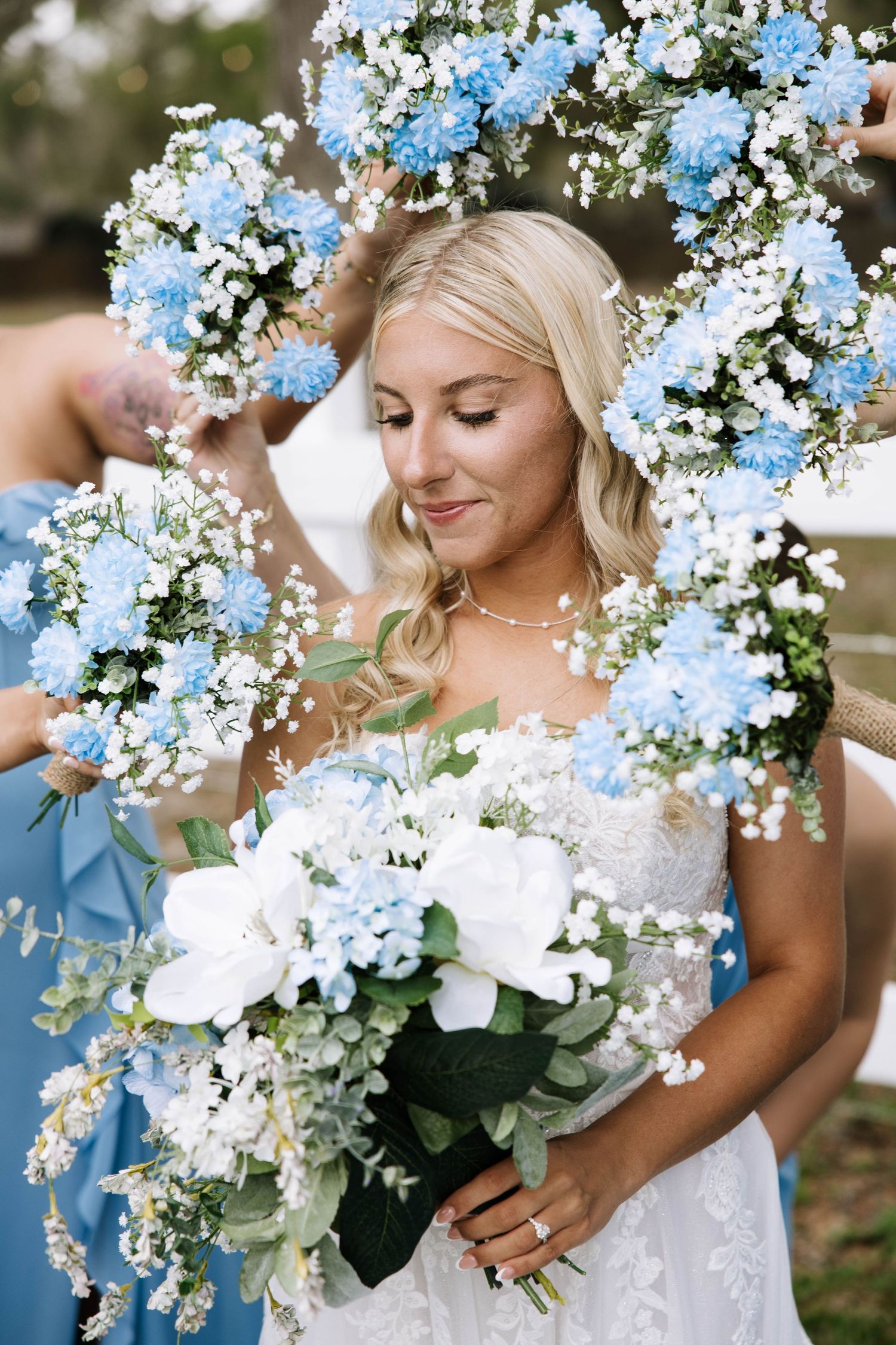 Blonde bride holds white and blue bouquet framed by matching floral wreath at wedding ceremony.
