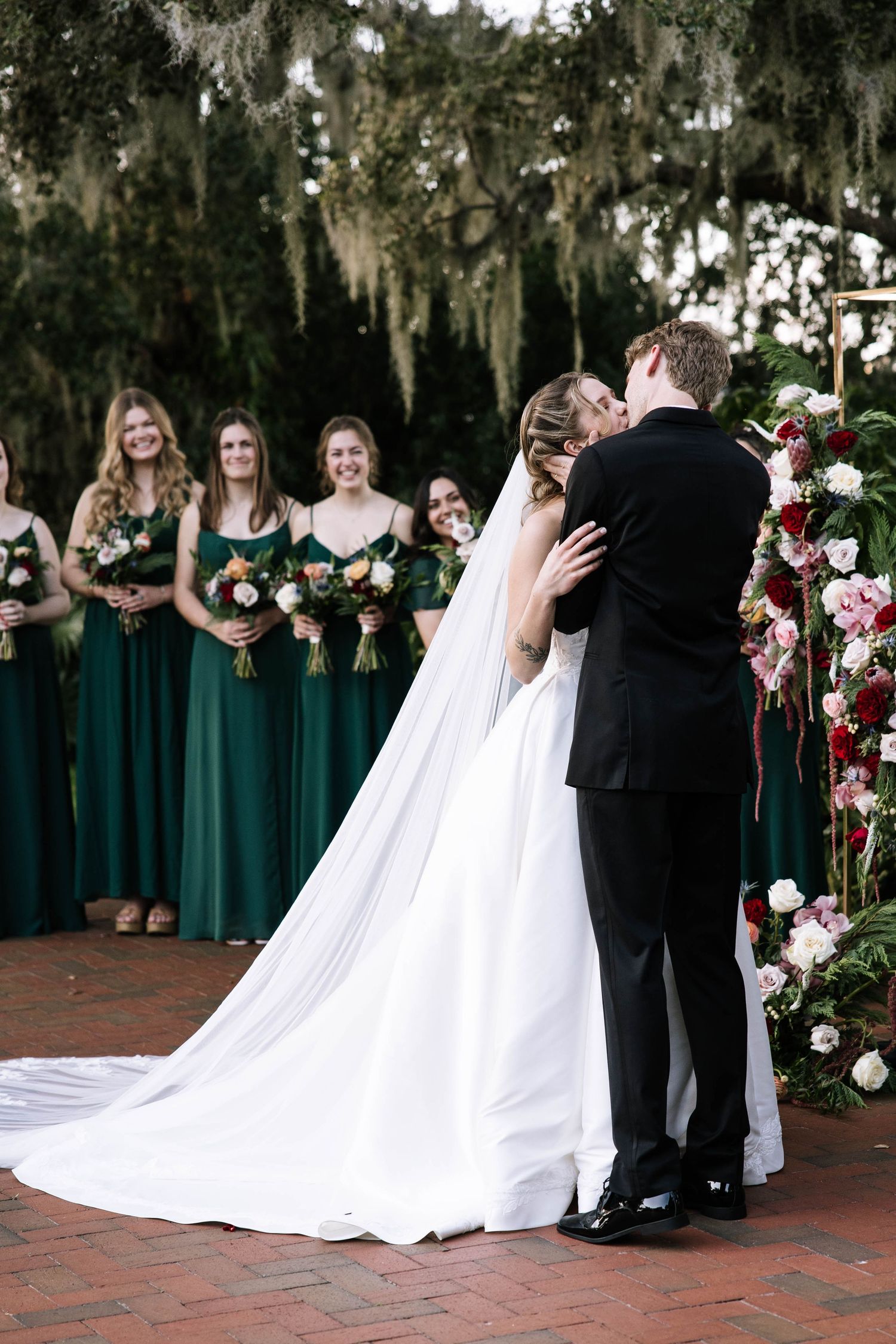 Bride and groom share first kiss at outdoor ceremony with bridesmaids in emerald green dresses under Spanish moss.