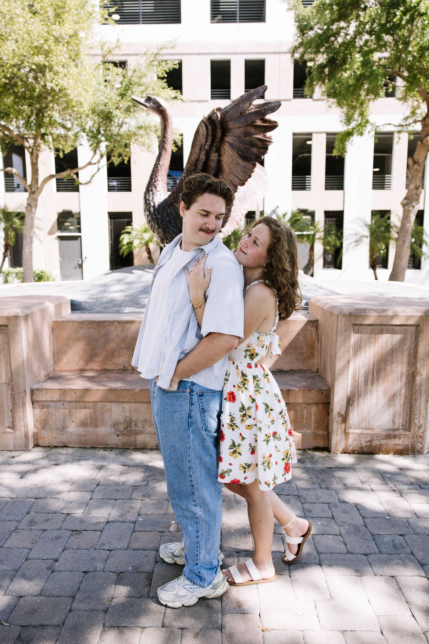 Couple poses playfully before winged sculpture in sunny urban courtyard setting.