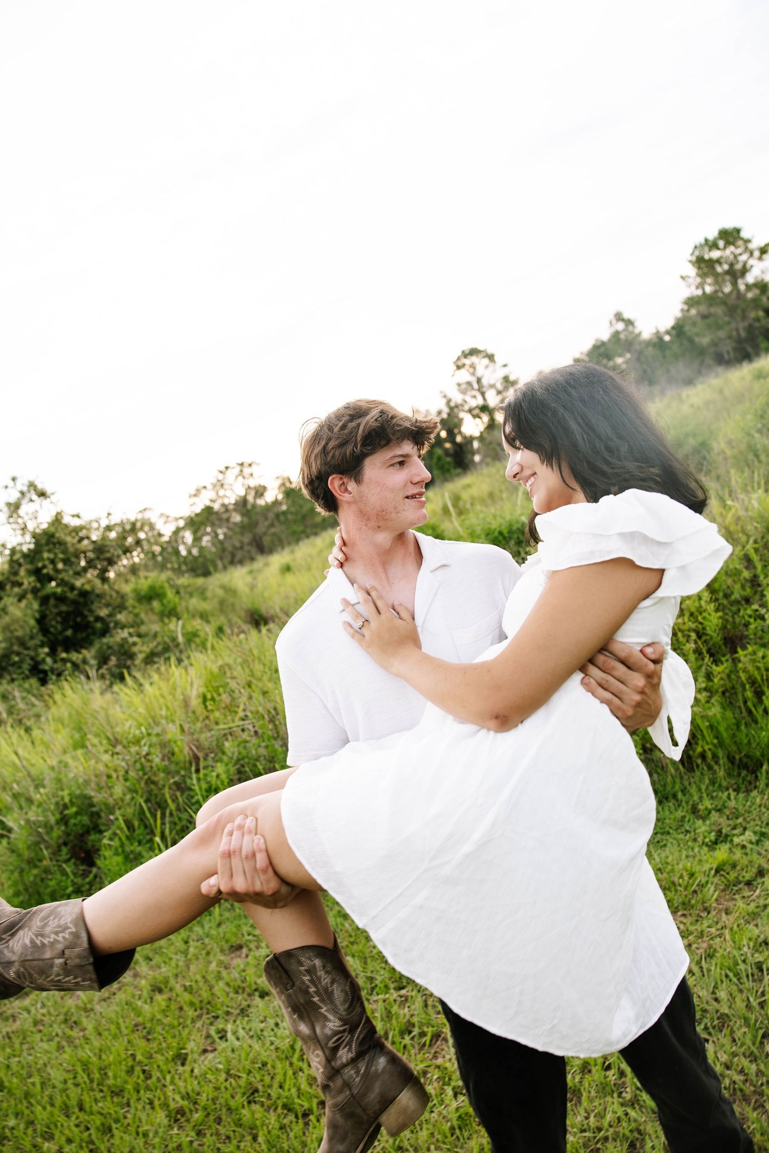 A man in white shirt lifts a smiling woman in white dress and cowboy boots in a lush green field.
