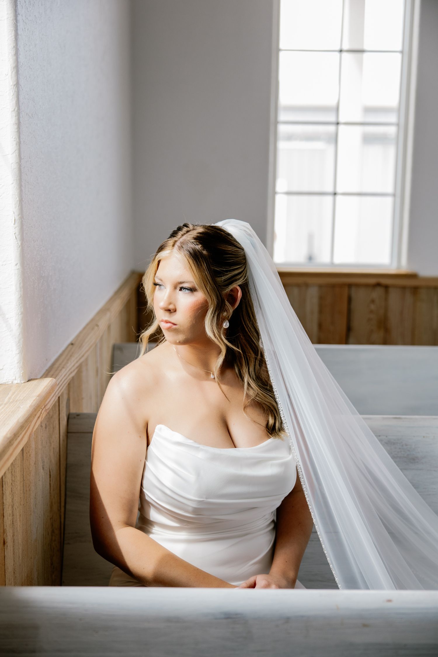 Beautiful bride in strapless white gown and cathedral veil seated in wooden church pew by bright window.