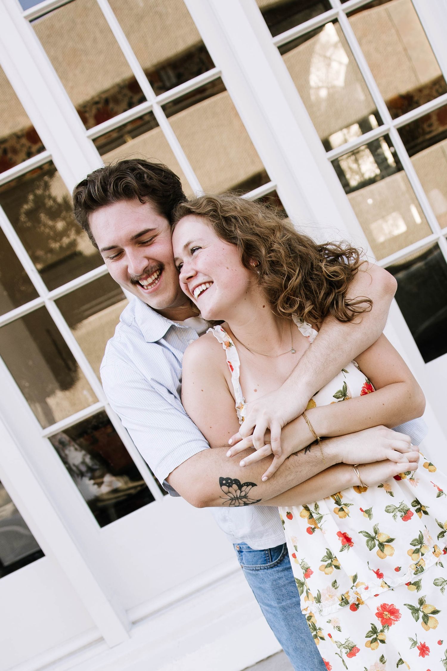 A laughing couple embraces joyfully in front of large white-framed windows, the woman in a floral dress.