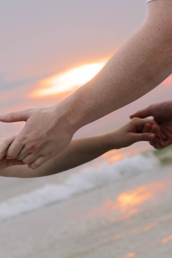 Close-up of two people gently holding hands at sunset on a beach with pink sky reflecting on water.
