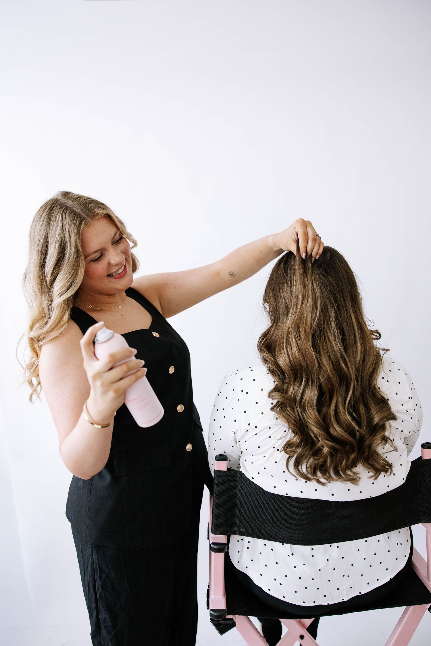 Blonde hairstylist in black dress sprays product on client with long wavy brunette hair.Photography Studio in Bartow Florida