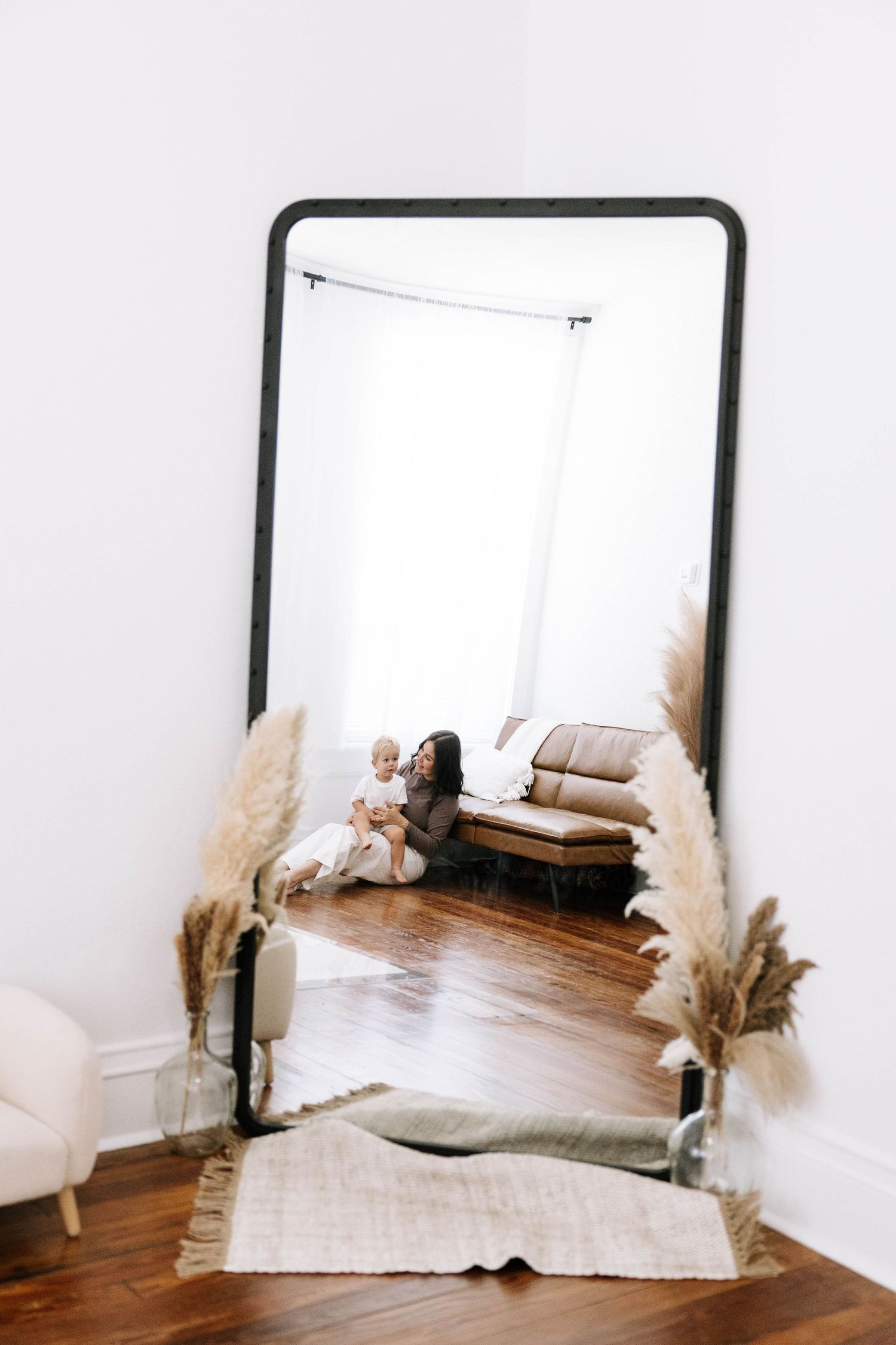 Large floor mirror reflecting mother sitting on hardwood floor holding blonde toddler near pampas grass decor. Photography Studio in Bartow