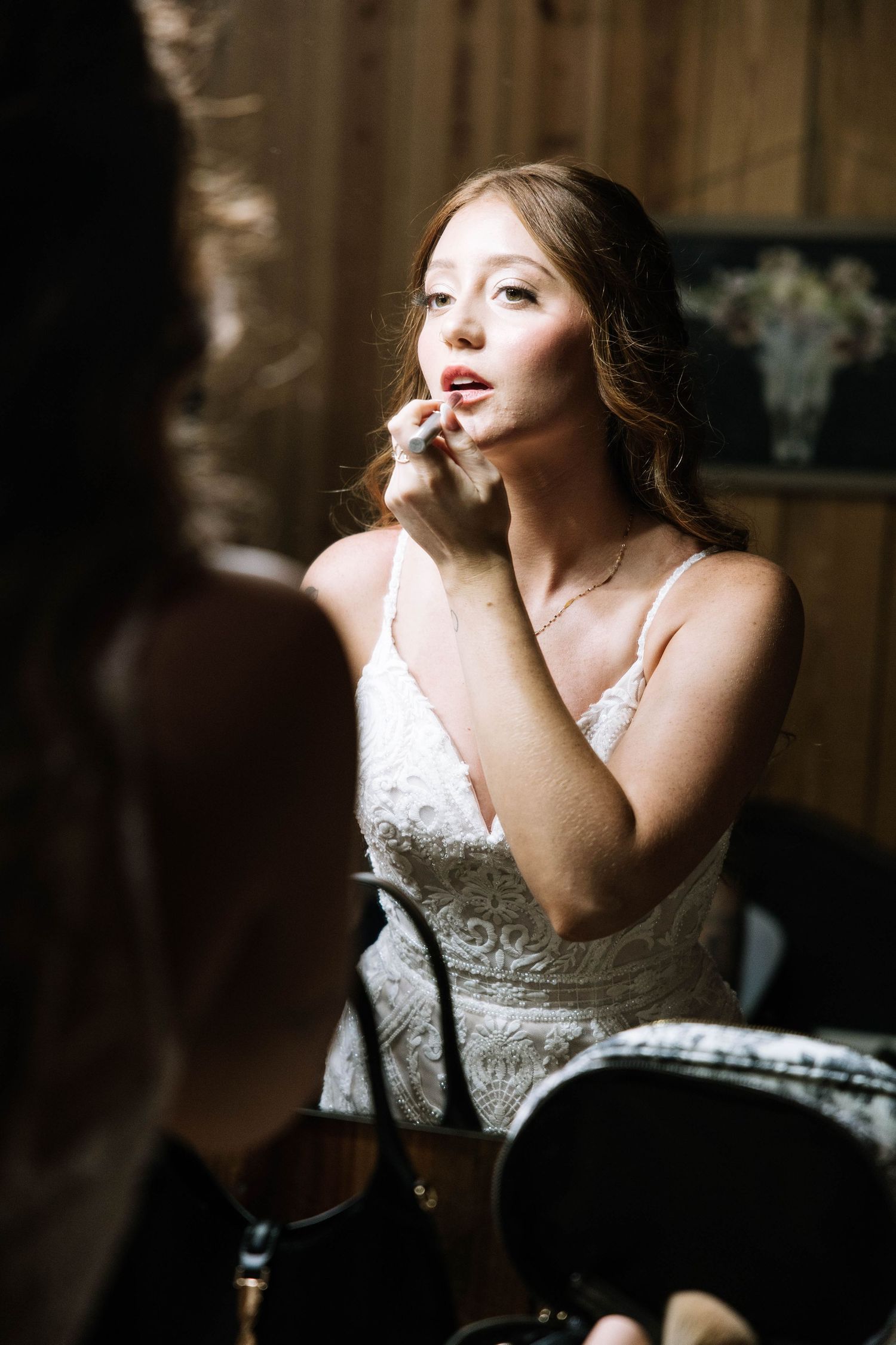 Bride in white lace gown applying lipstick while seated before a mirror in a warmly lit rustic setting.