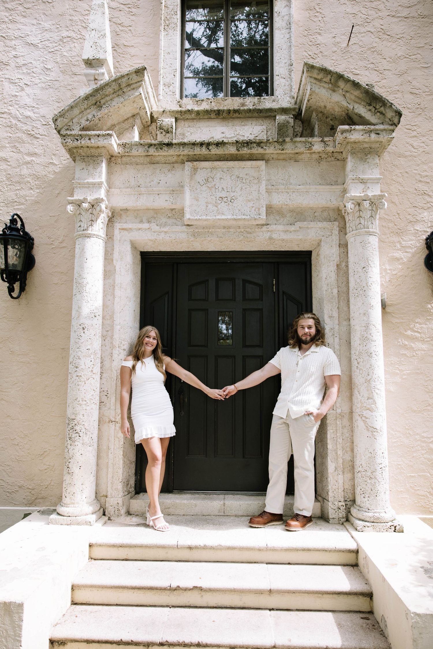 Couple in white attire holds hands on steps of elegant stone building entrance with ornate doorway.