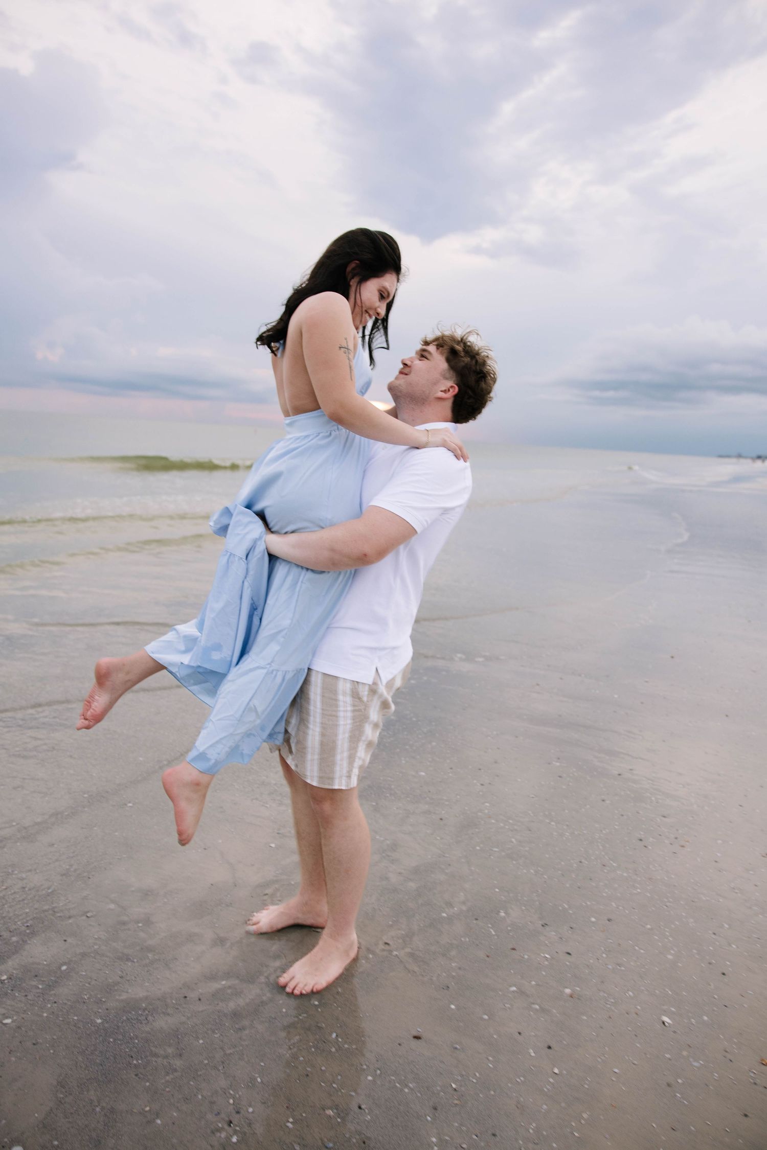 Man lifting woman in blue dress on a beach under a cloudy sky.
