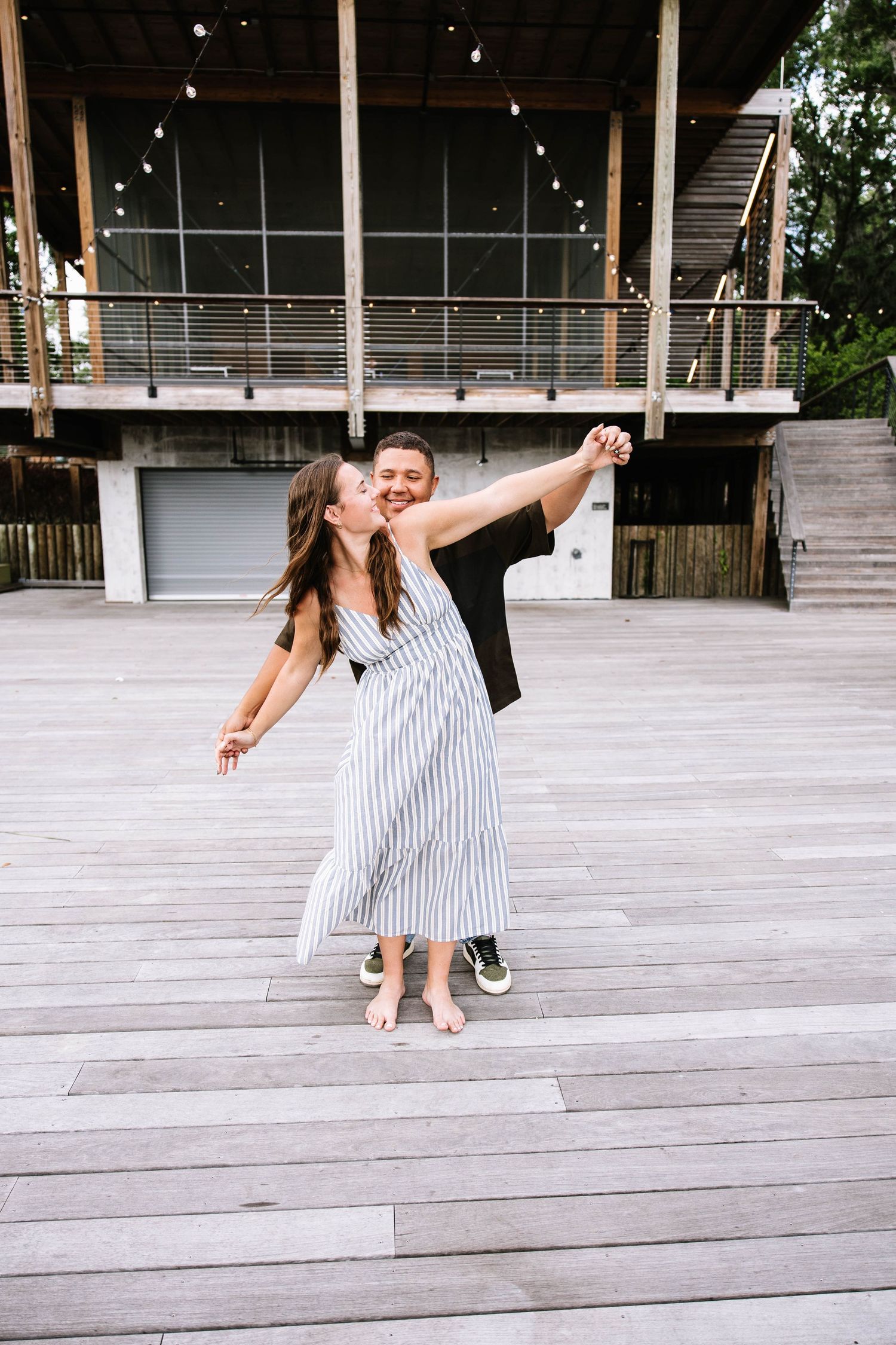 A happy couple dances playfully on an outdoor wooden stage with string lights overhead.