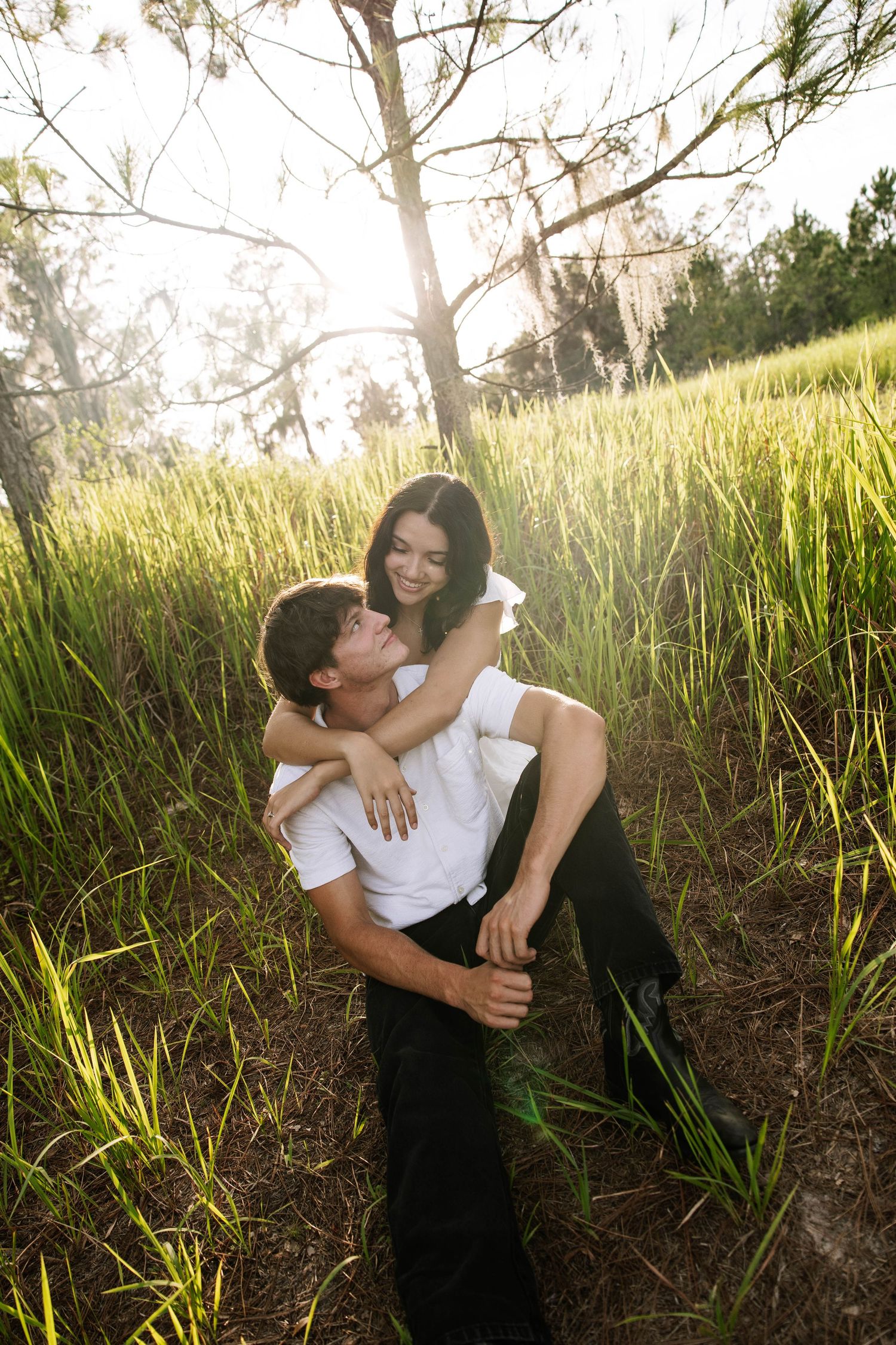 A couple embraces in a sunlit green field, the man lifting the smiling woman in a romantic outdoor setting.
