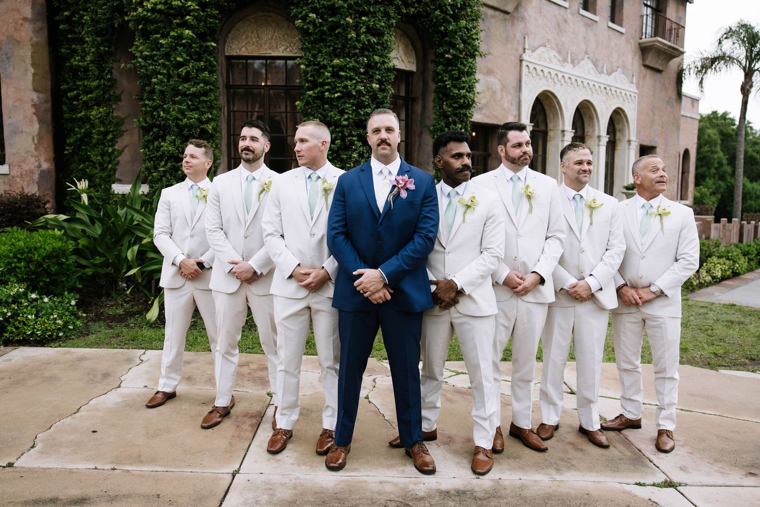 Groom in navy blue suit stands with groomsmen in white suits and pink boutonnieres outside elegant venue.