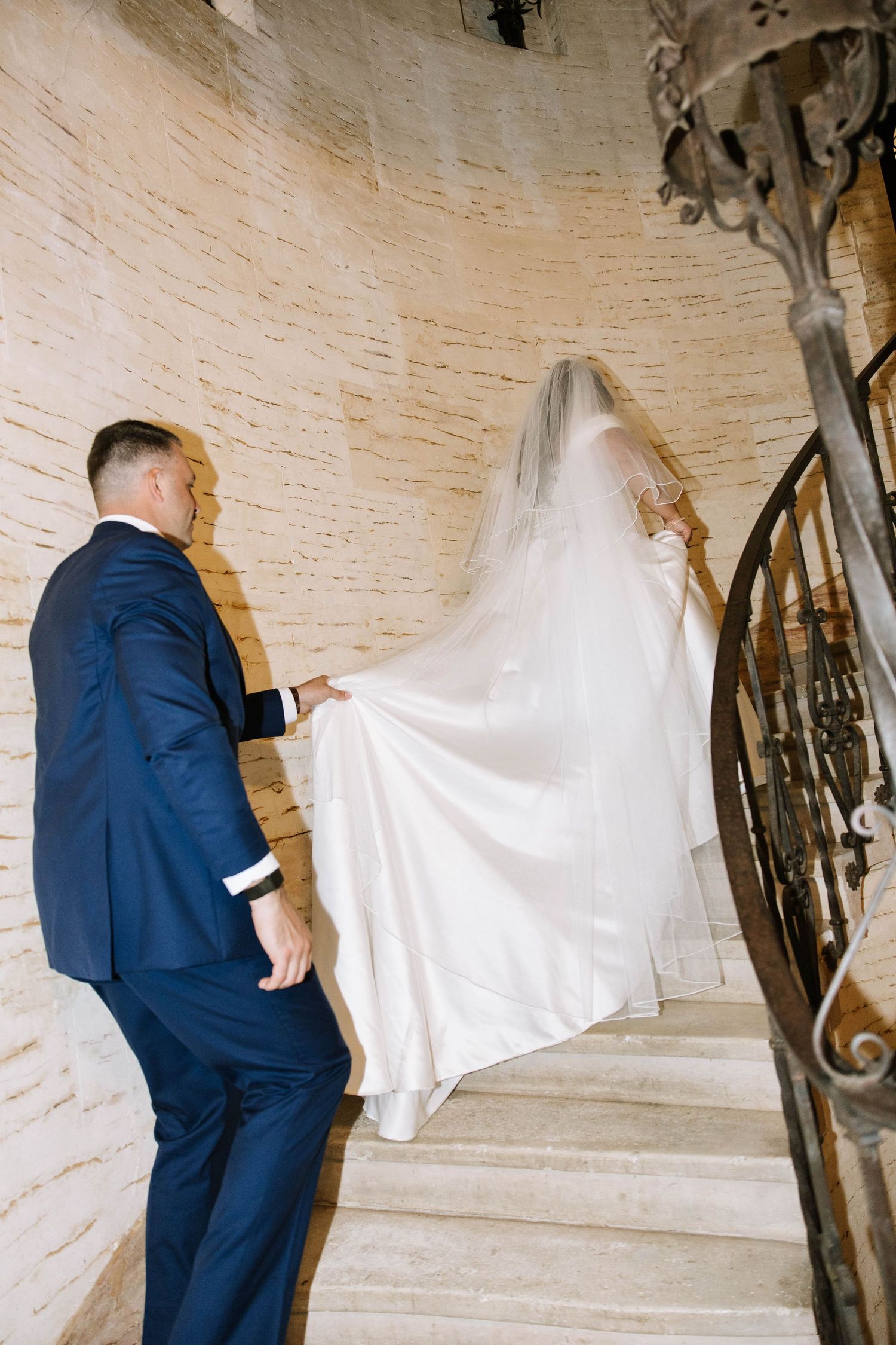Groom in navy suit holds bride's white gown train as couple ascends elegant stone spiral staircase.