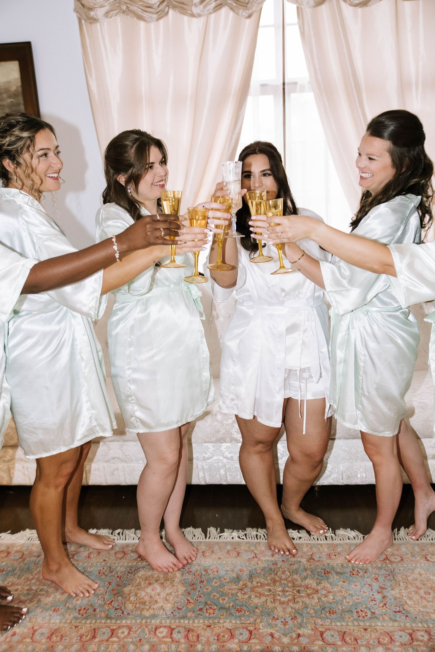 Four bridesmaids in satin robes toast champagne glasses together while getting ready for wedding.