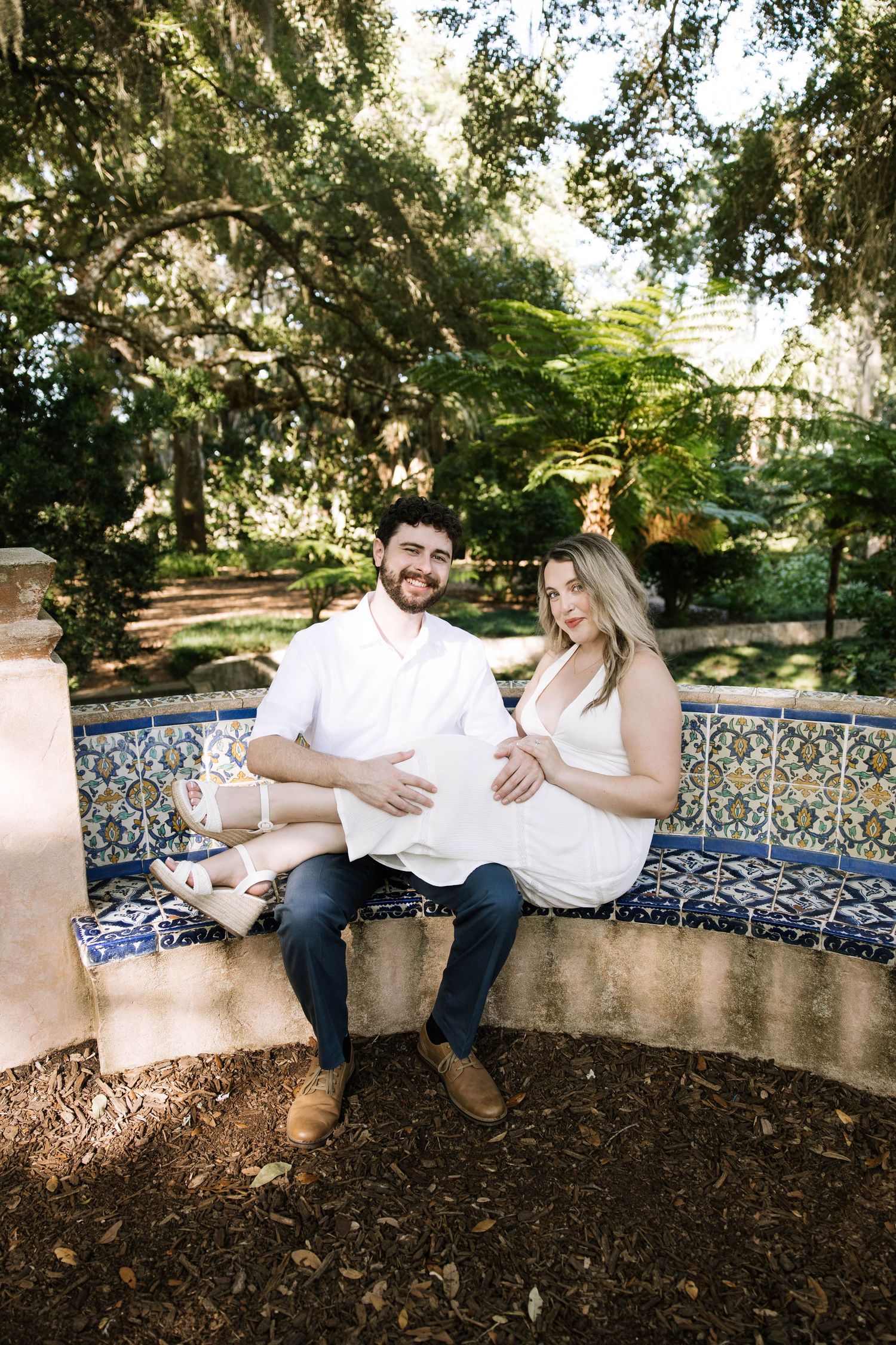 Couple sitting on mosaic tile bench in shaded garden during maternity session.