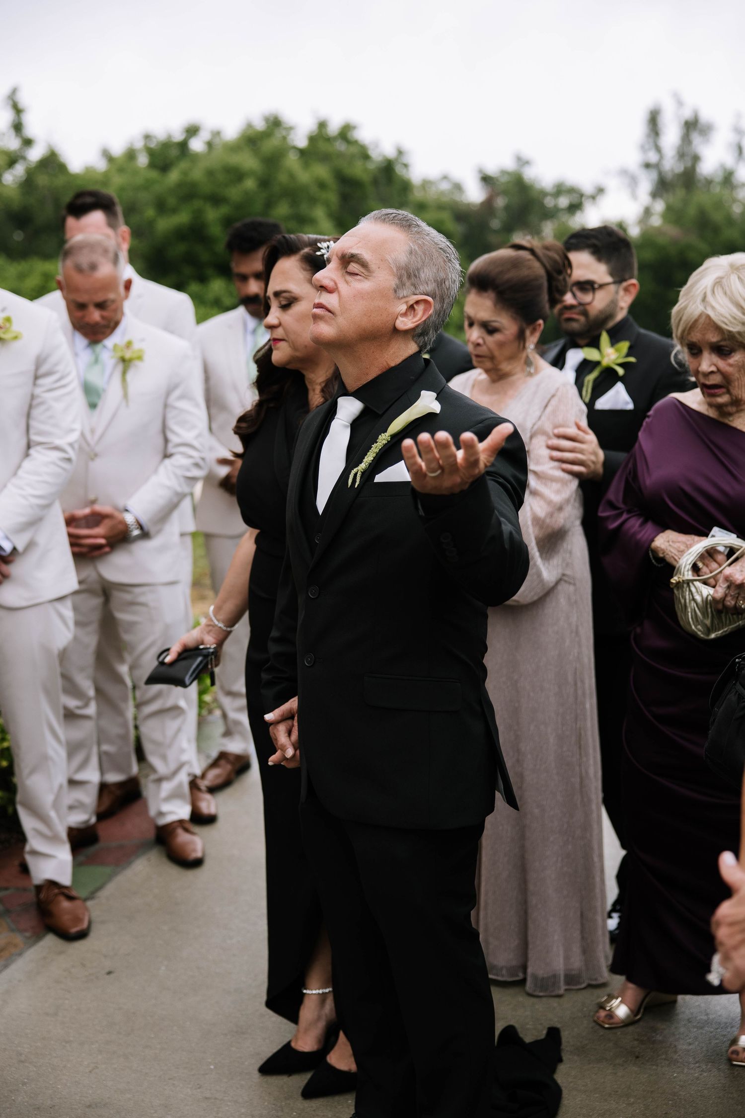 Man in black tuxedo blowing a kiss at an outdoor wedding ceremony with guests dressed in formal attire.