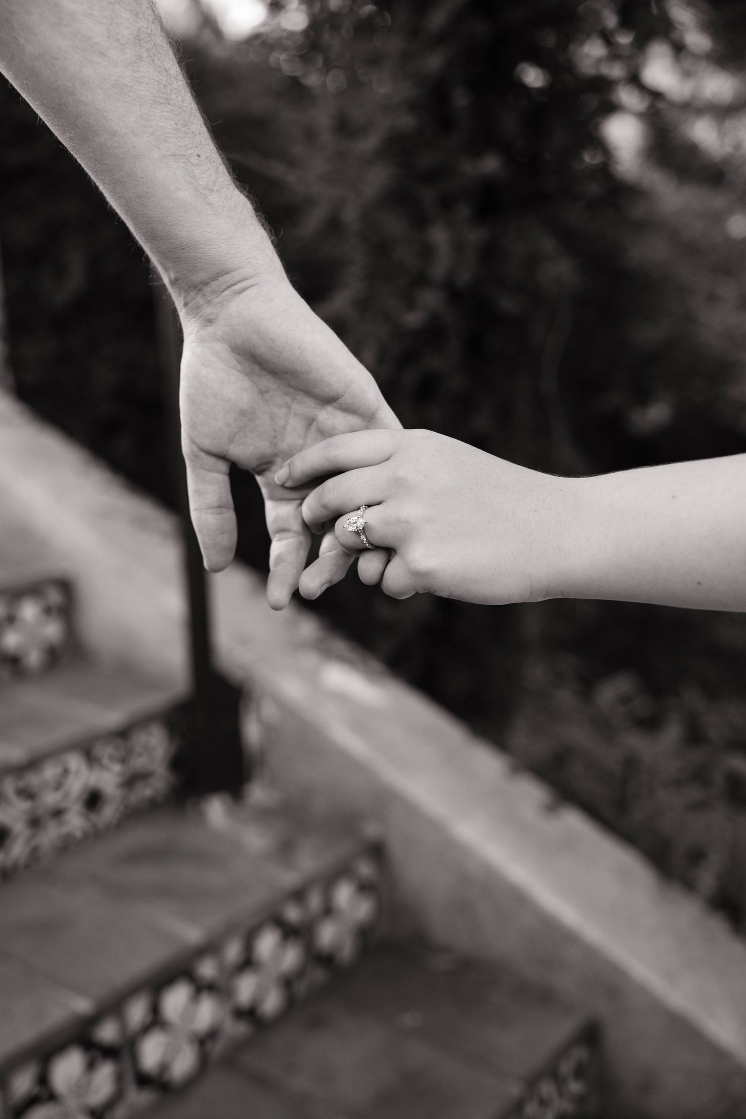 A close-up black and white photo of a couple gently holding hands near decorative tiled steps outdoors.