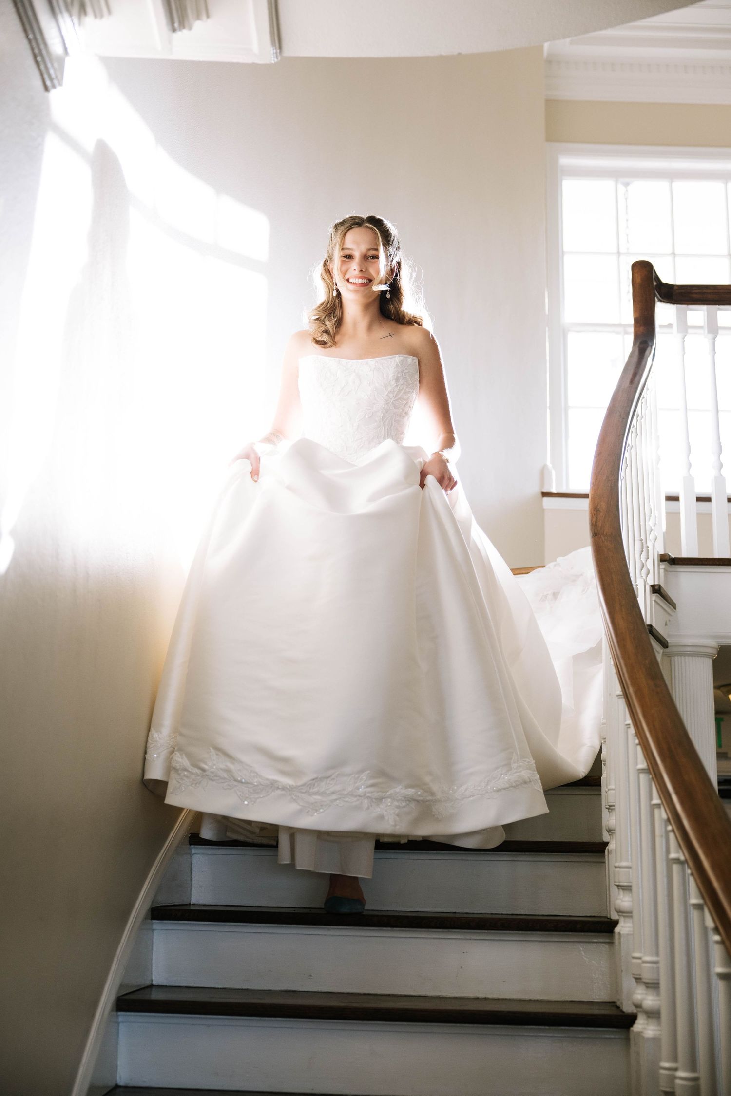 Smiling bride in strapless ball gown and veil descending a sunlit staircase.