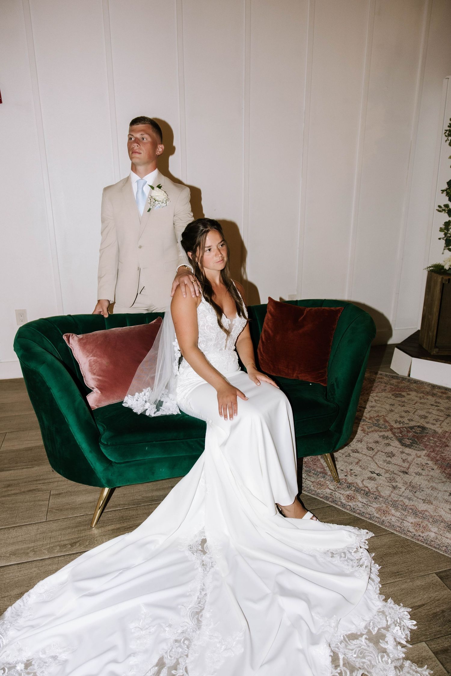 Bride in white lace gown sits on green velvet sofa while groom in beige suit stands behind her.