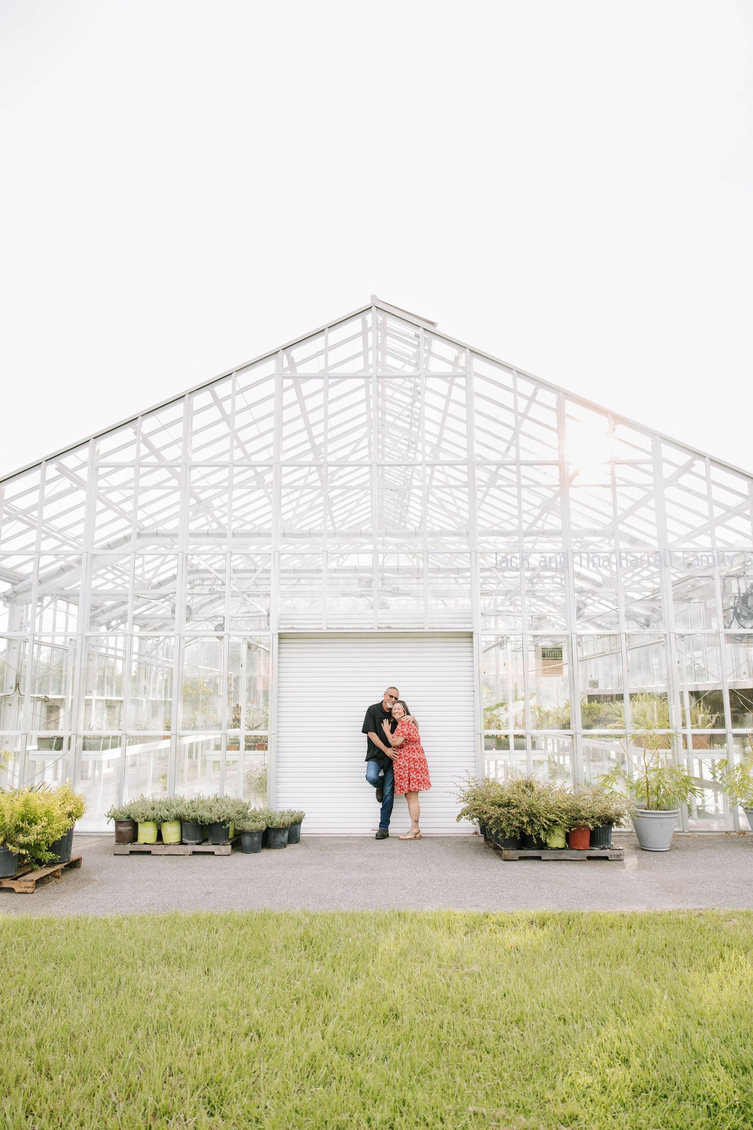 Couple embracing in front of a large white greenhouse with potted plants nearby.
