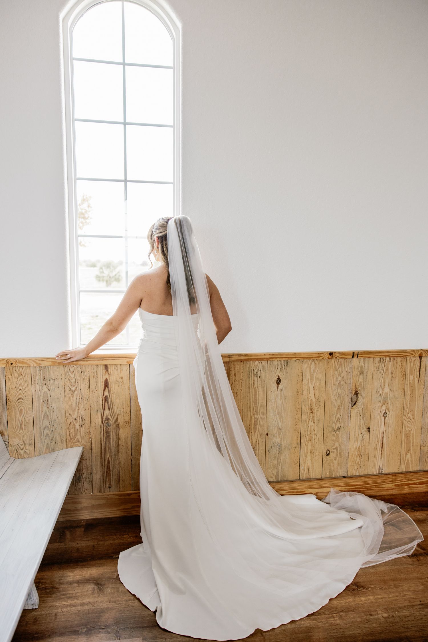Bride in fitted white gown with cathedral veil stands looking out tall arched window in chapel.