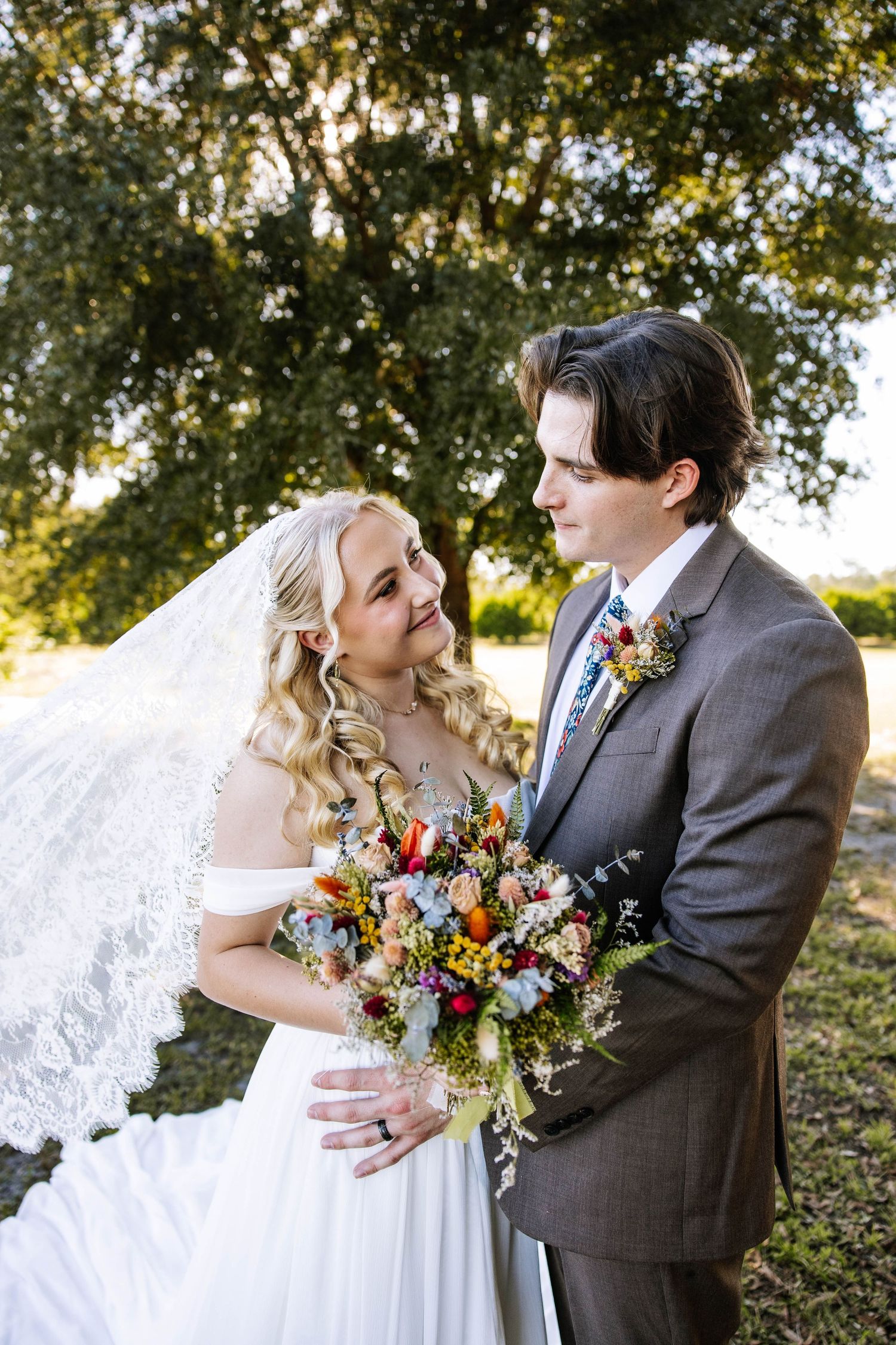 Bride and groom share a tender moment outdoors, bride holding colorful wildflower bouquet with lace veil flowing in the breeze.