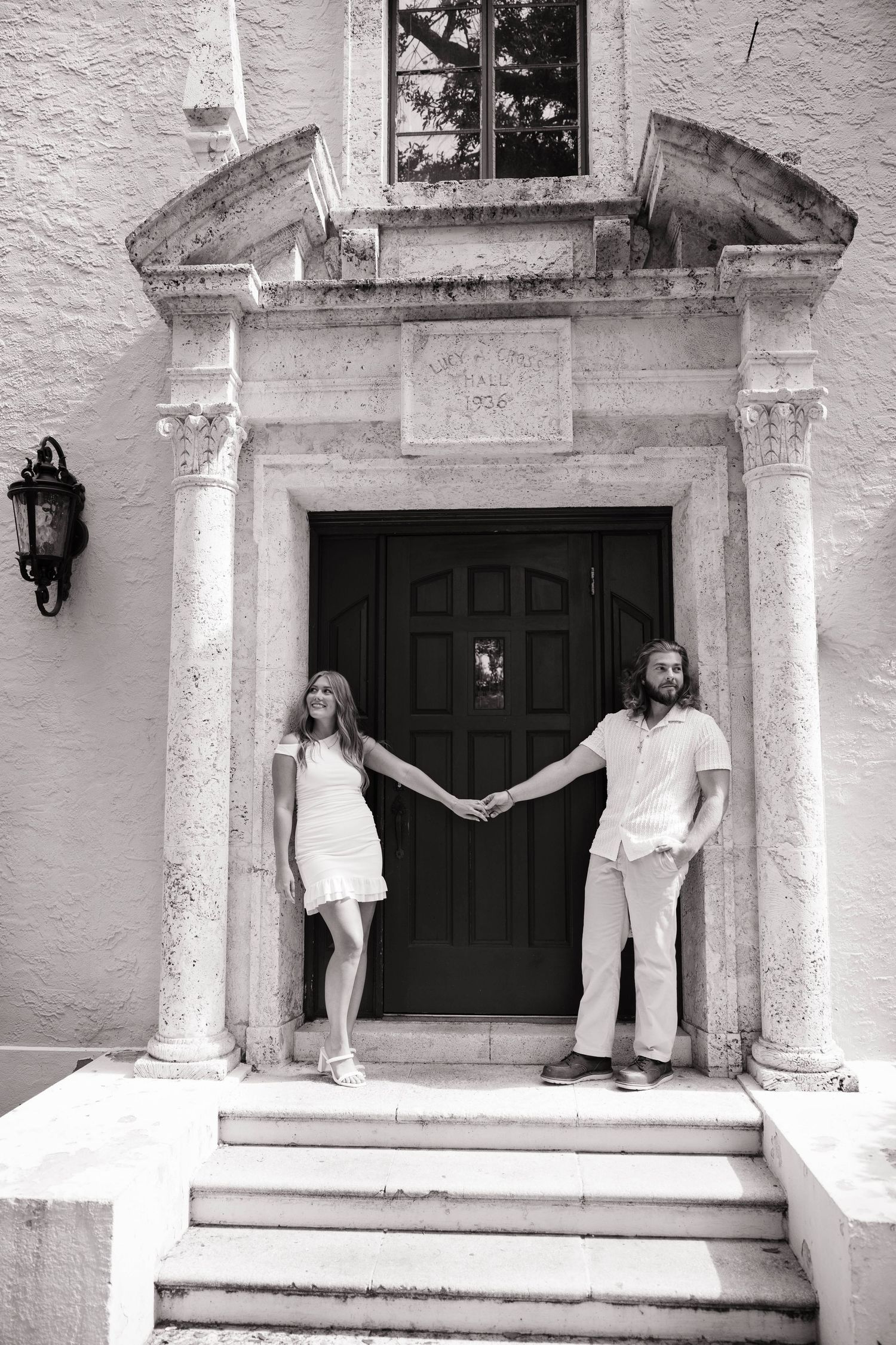 A black and white photo of a couple holding hands while standing at the entrance of a historic stone building.