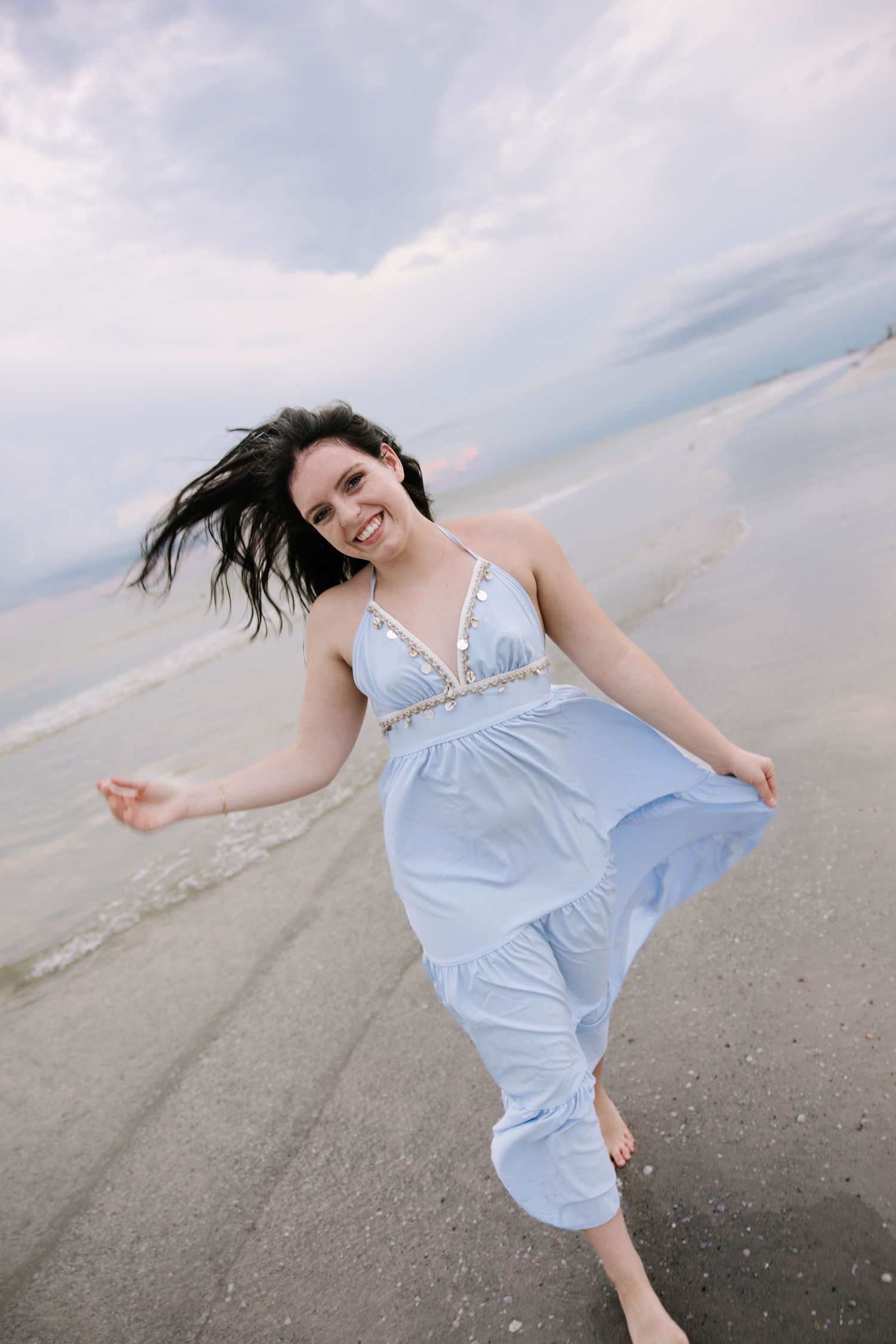 A joyful dark-haired woman in a light blue maxi dress twirls barefoot on a sandy beach at dusk.