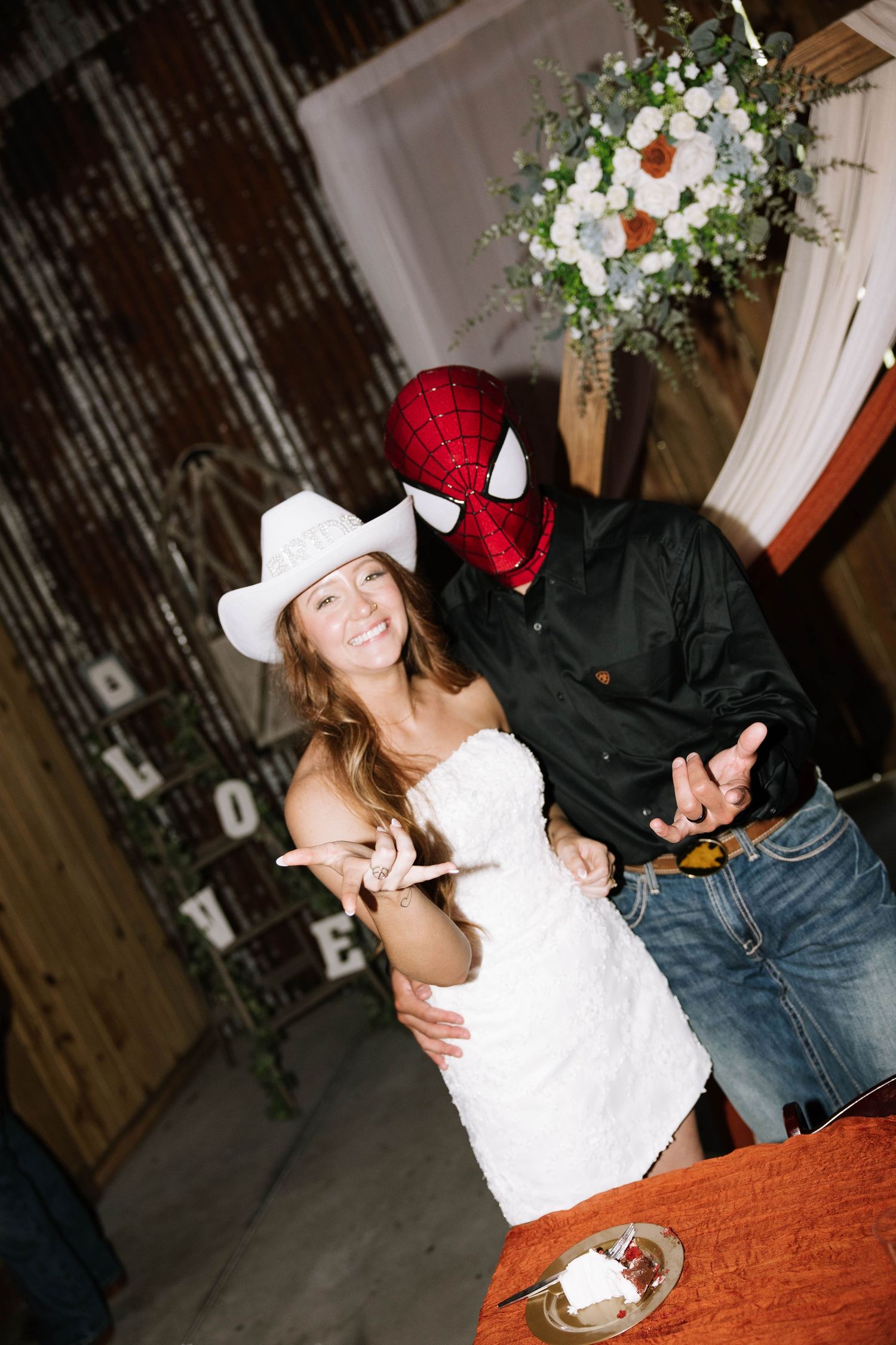 Smiling bride in white cowboy hat poses with groom wearing a Spiderman mask at rustic barn wedding.