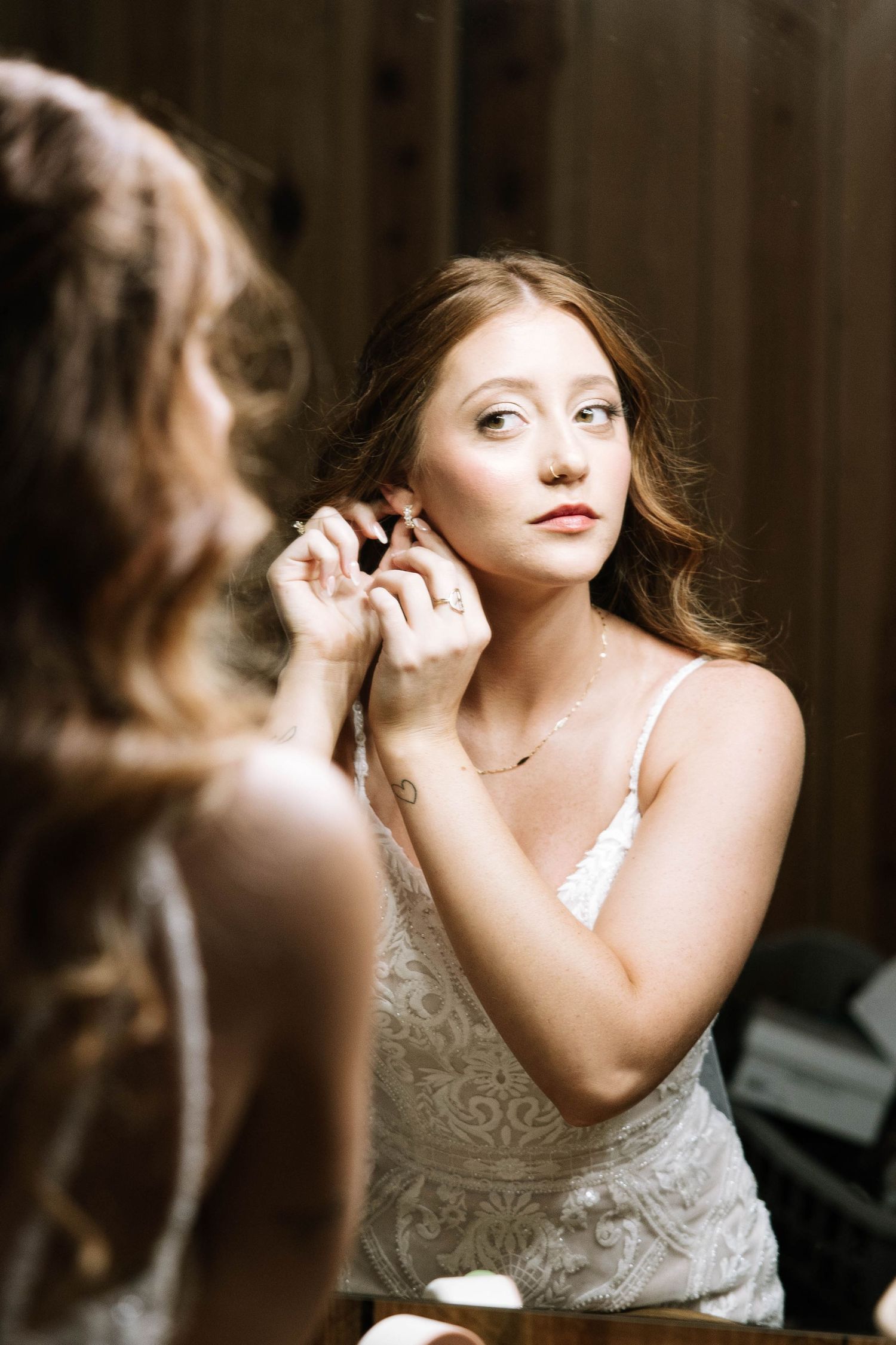 Bride putting on earring while looking in mirror during wedding day preparations.