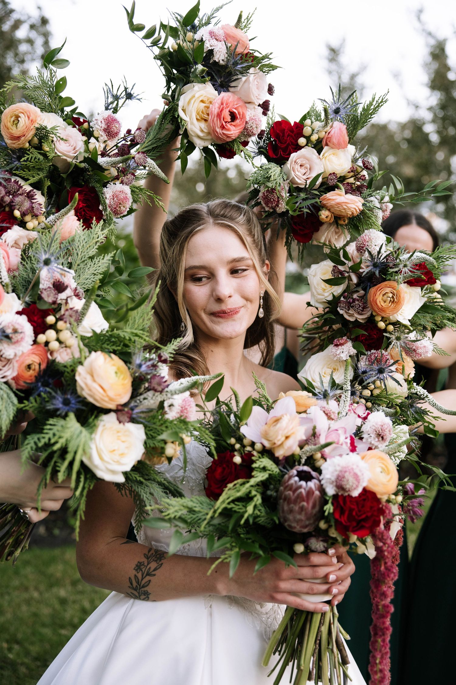 Smiling bride in white gown surrounded by colorful bridesmaids bouquets featuring roses, protea, and wildflowers.