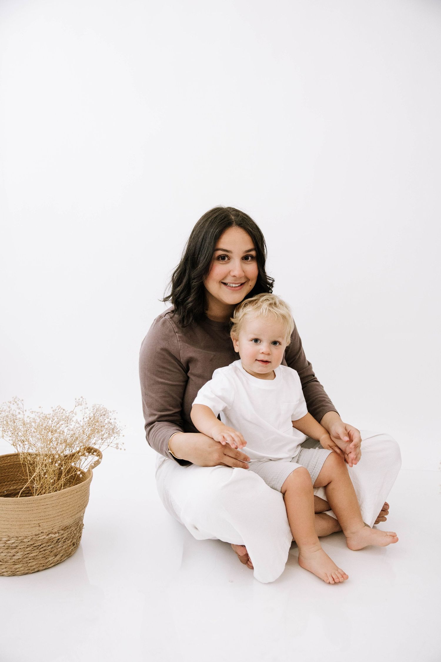 Smiling dark-haired mother in brown top sitting on white floor holding blonde toddler beside woven basket. Photography Studio in Bartow