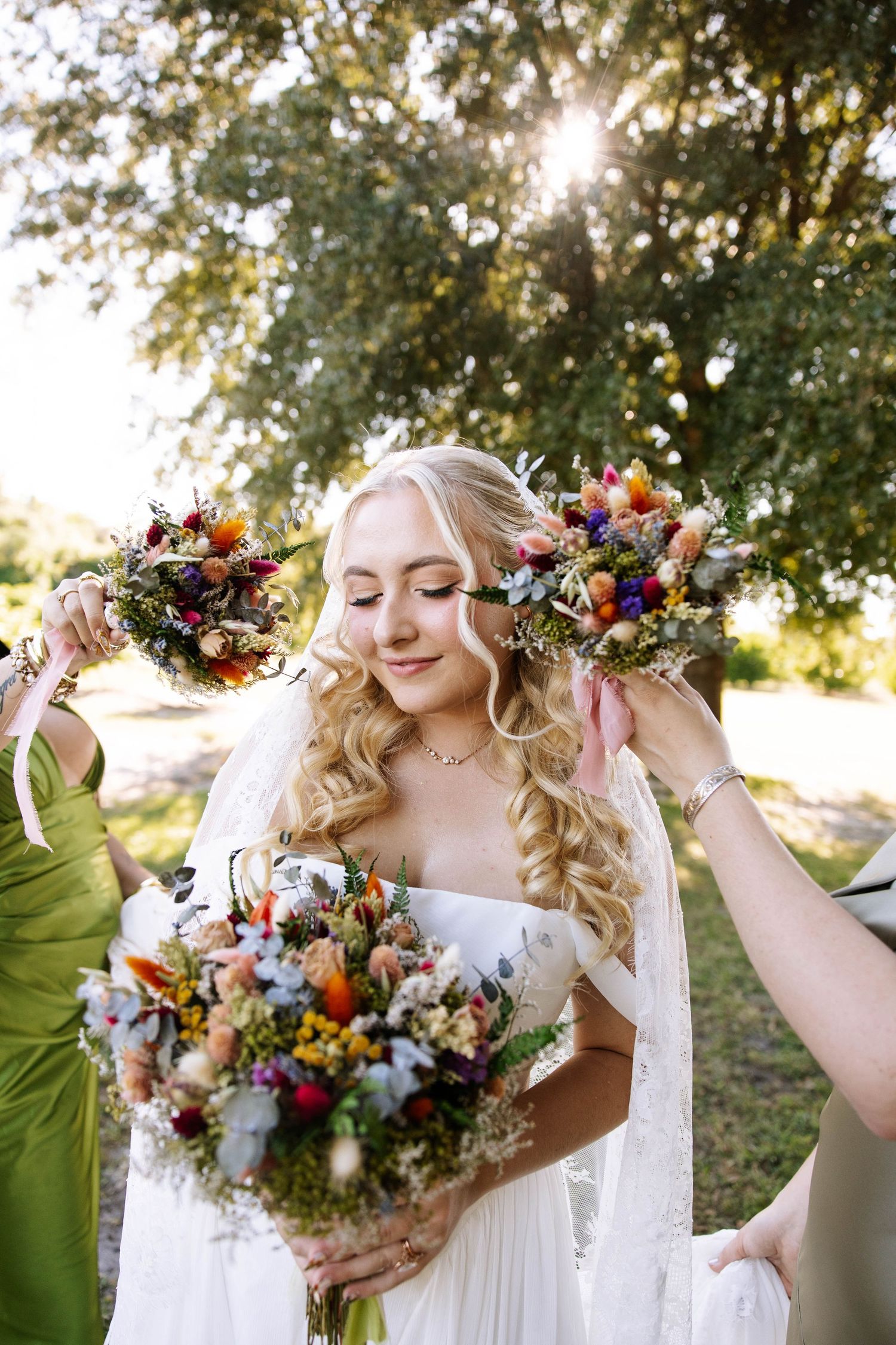 Smiling bride in white gown surrounded by colorful wildflower bouquets held by bridesmaids outdoors in golden sunlight.