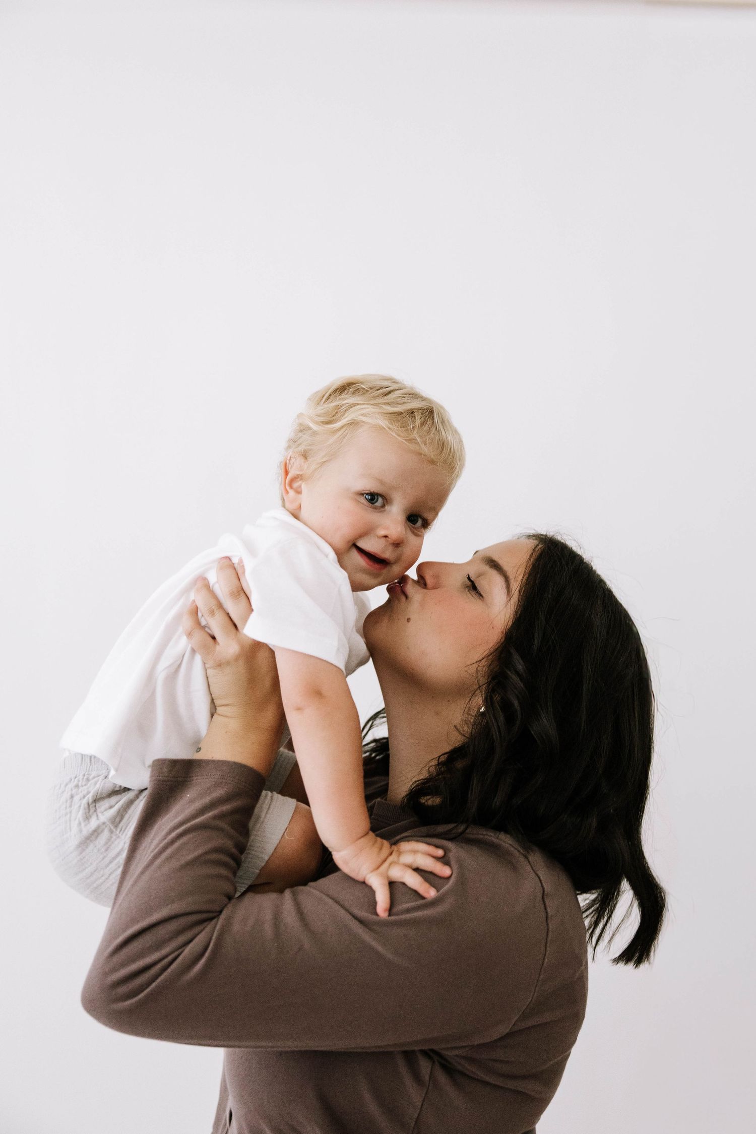 A mother lovingly kisses her smiling blonde toddler while lifting the baby up against a white background.