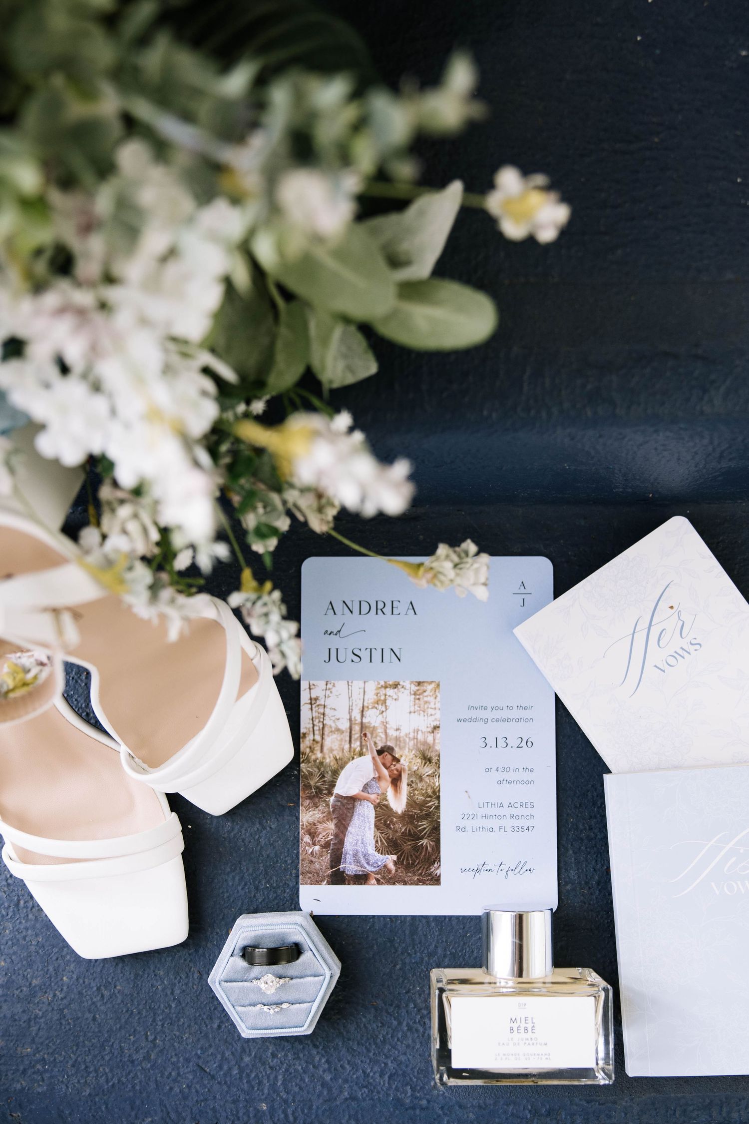 Wedding flat lay featuring invitation suite, bridal heels, engagement ring, and perfume on navy background.