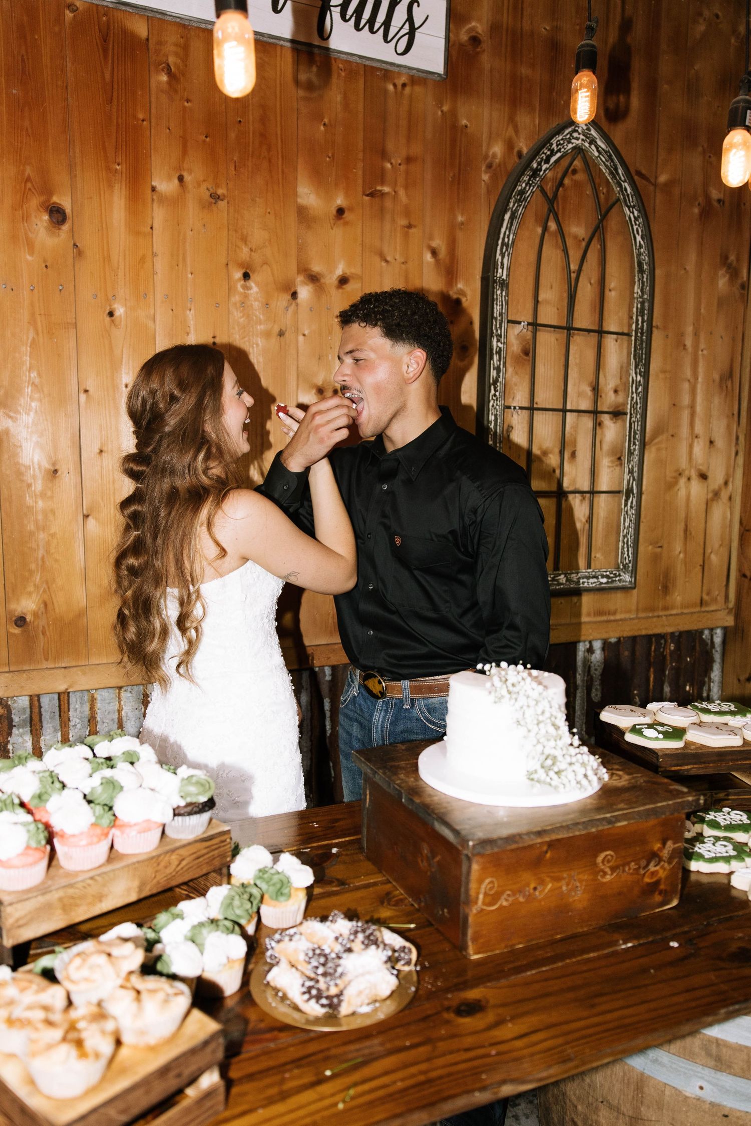 Bride feeds groom cake at rustic wooden dessert table with cupcakes.