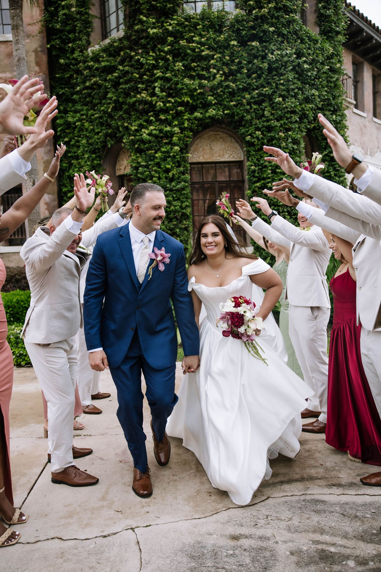 Bride and groom walk through a tunnel of raised hands from wedding party outside an ivy-covered venue.