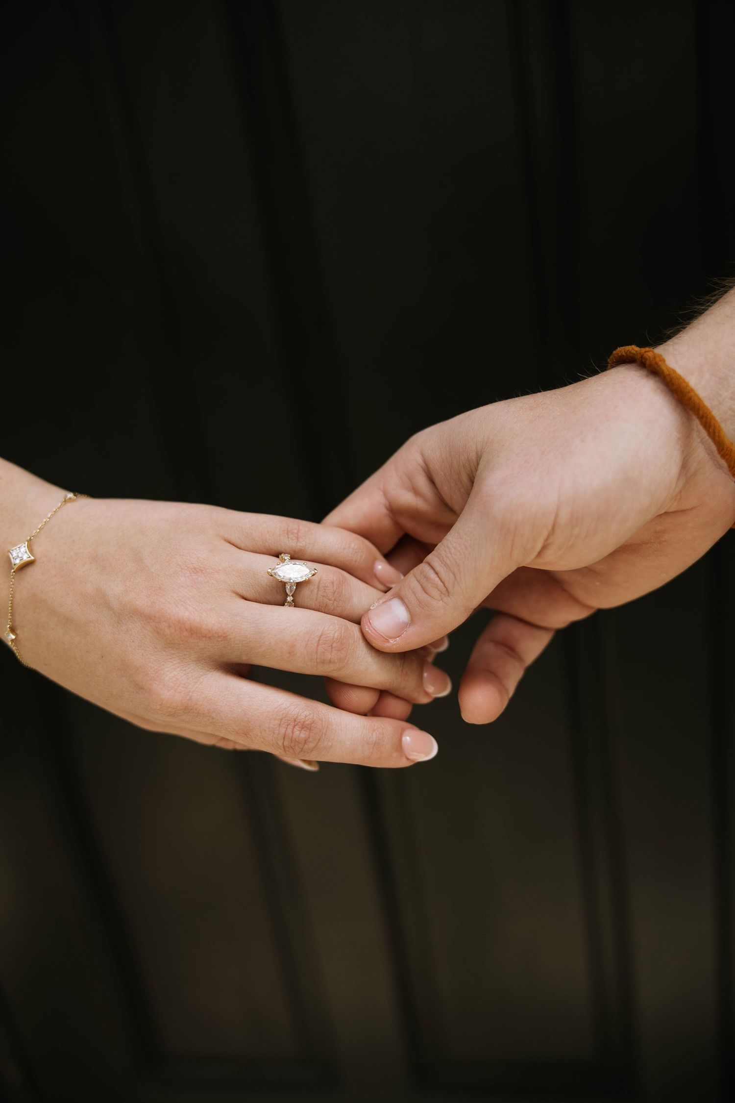 Close-up of couple holding hands showing elegant engagement ring and gold bracelet on dark background. Rollins College.