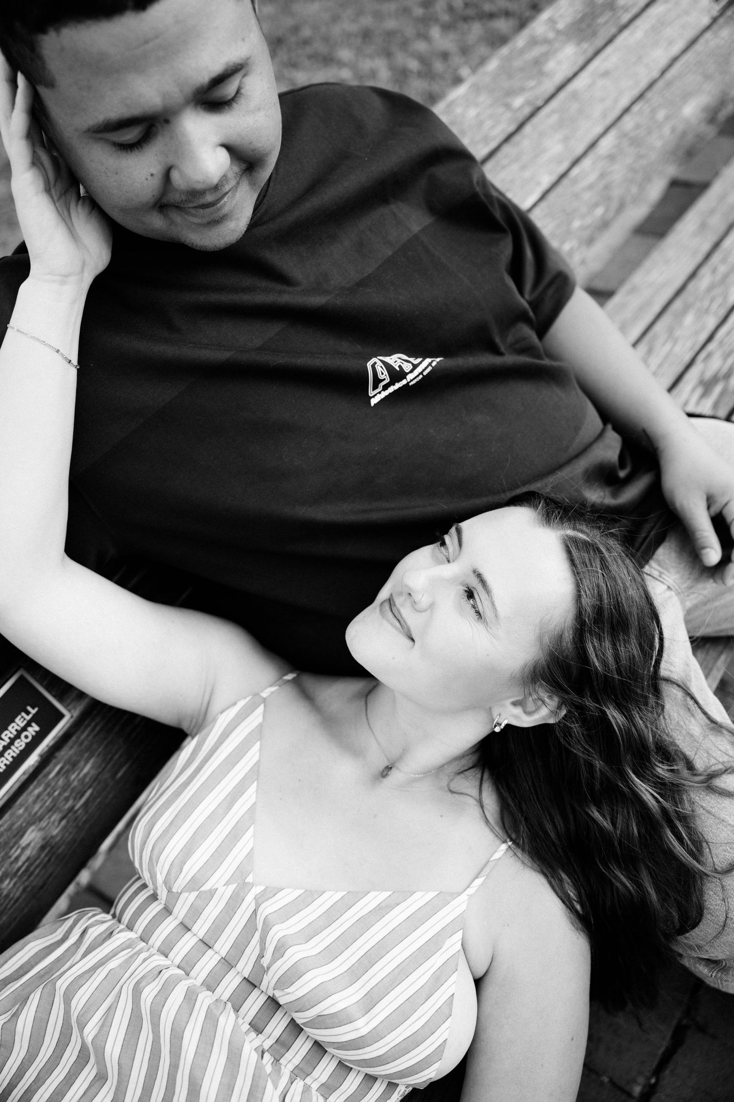 A black and white photo of a couple relaxing on a wooden bench, gazing at each other affectionately.